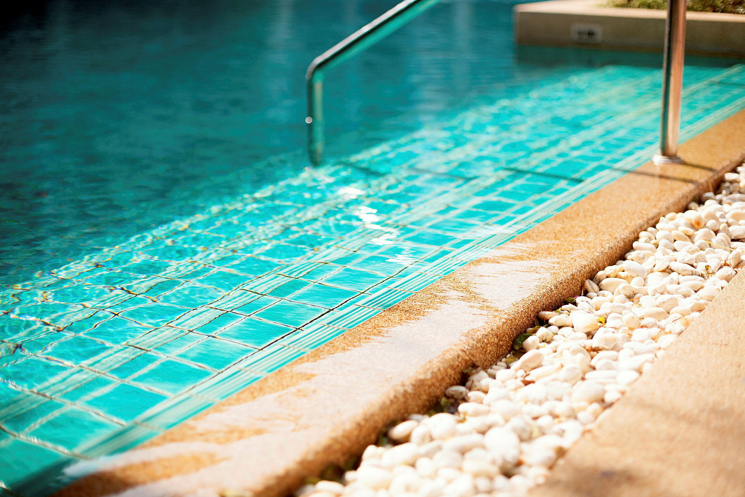 Nærbillede af kanten af en swimmingpool med blå fliser. Detaljer af vandoverfladen, en metalstang samt hvide dekorationssten er synlige. (AI-genereret tekst)
