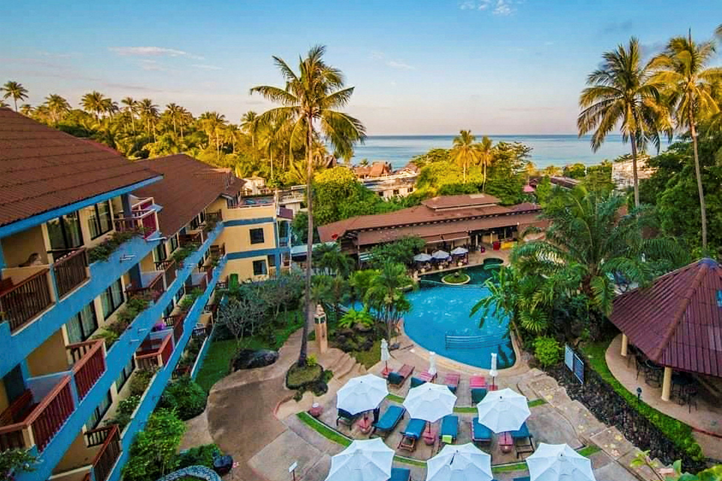 Udsigt over en hotelpool og have omgivet af bygninger og palmer, med havet i baggrunden. (AI-genereret tekst)