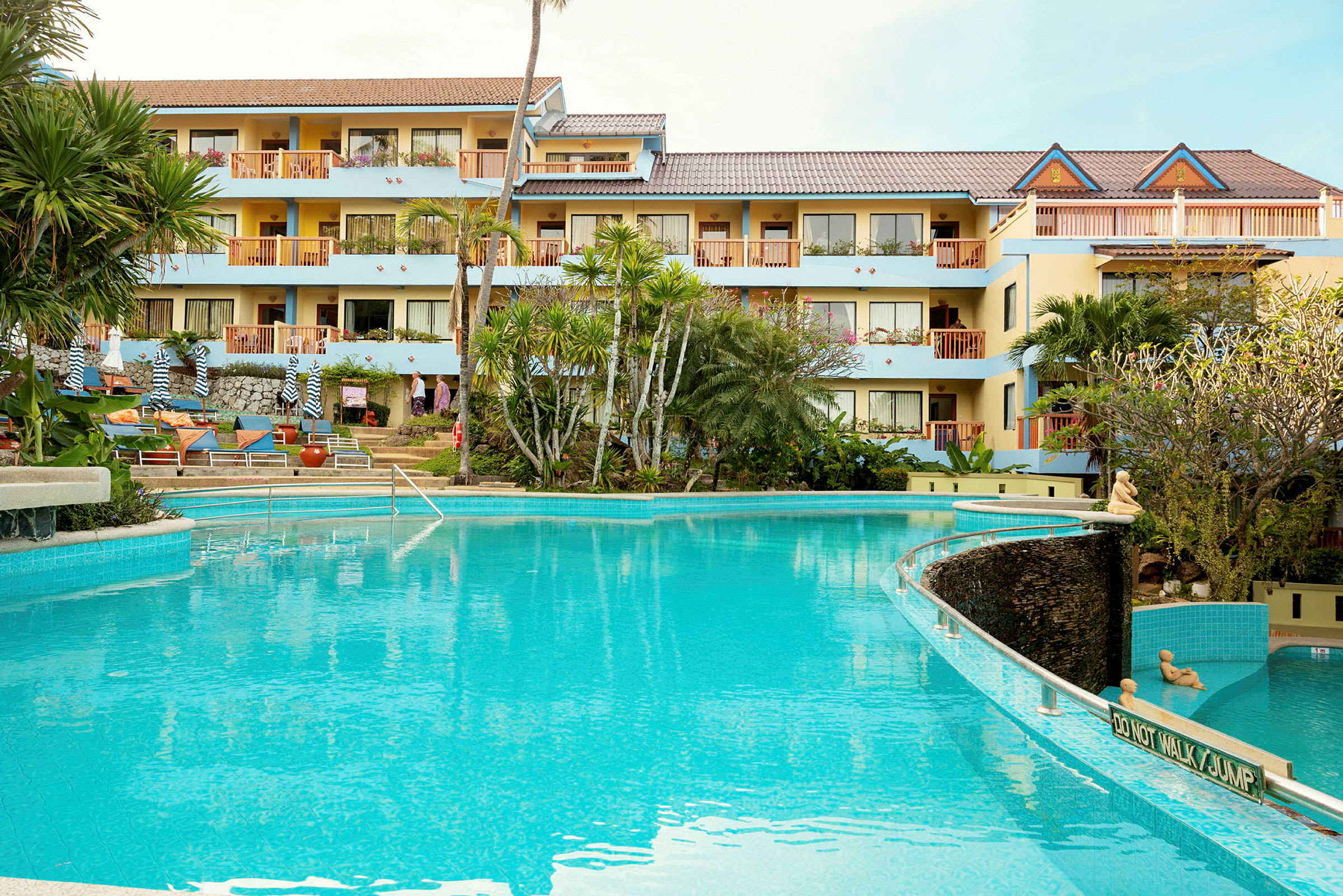 Hotelbygning med flere etager synlig bag en klarblå swimmingpool, omgivet af tropiske planter. (AI-genereret tekst)