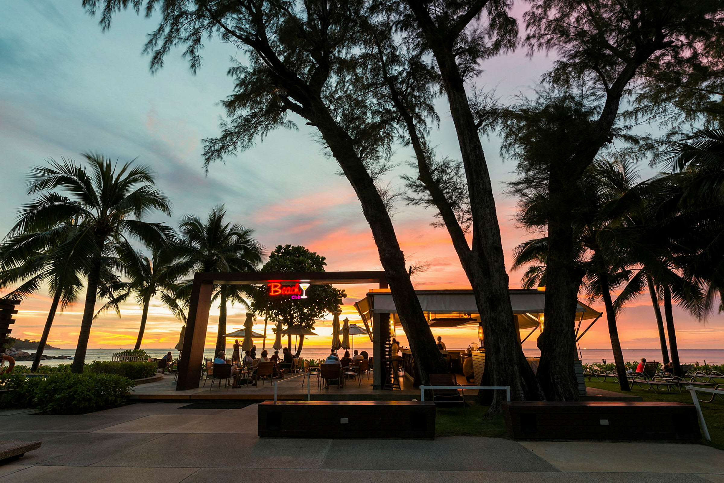 En strandbar med udsigt over havet ved solnedgang, omgivet af palmer. (AI-genereret tekst)