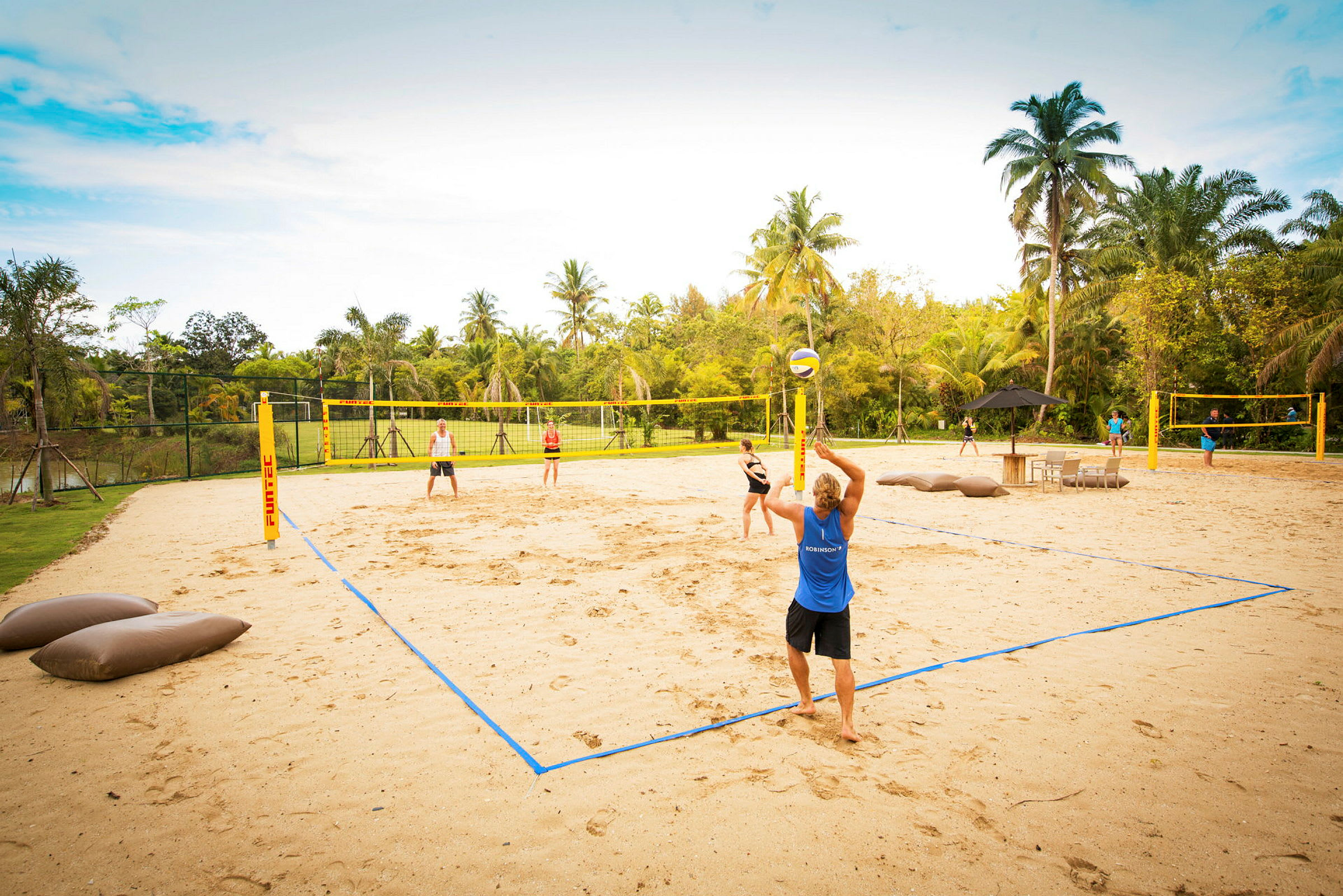 Flere personer spiller beachvolley på en sandbane omgivet af palmer og grønt område. (AI-genereret tekst)