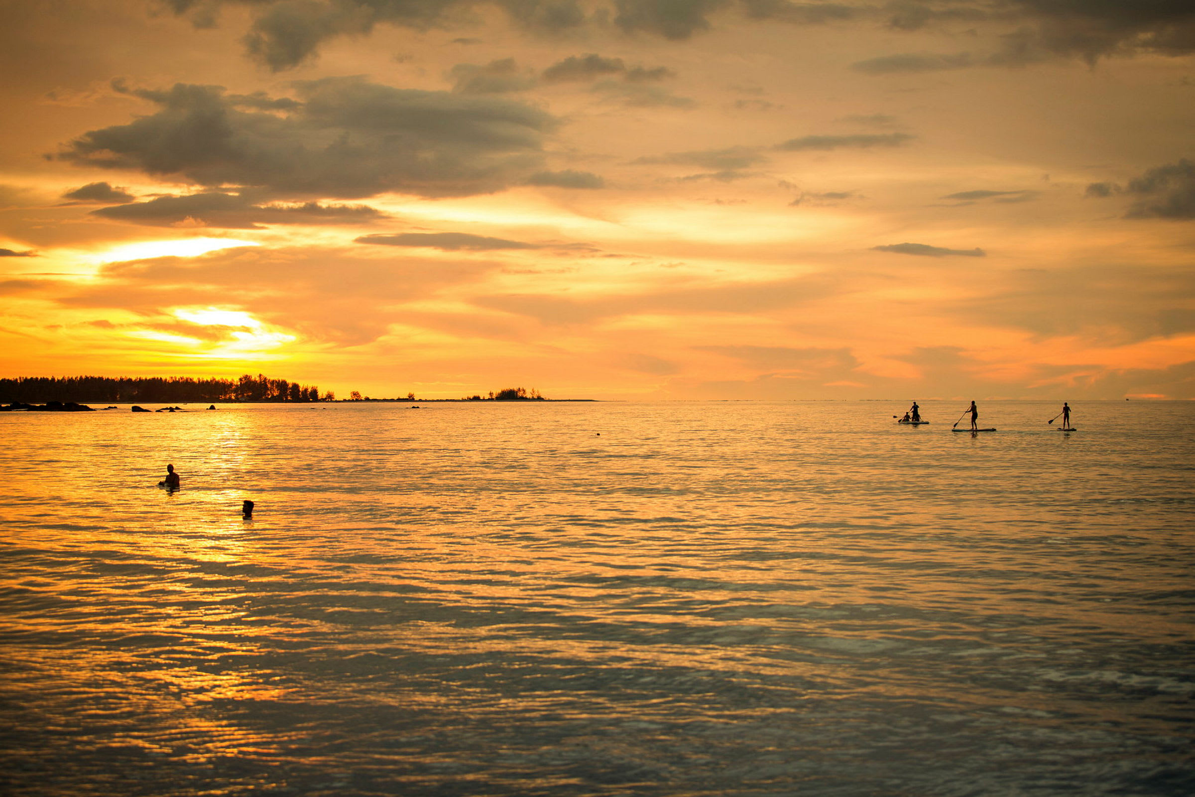 Personer padler på paddleboards i havet ved solnedgang, mens nogle andre svømmer i nærheden. (AI-genereret tekst)