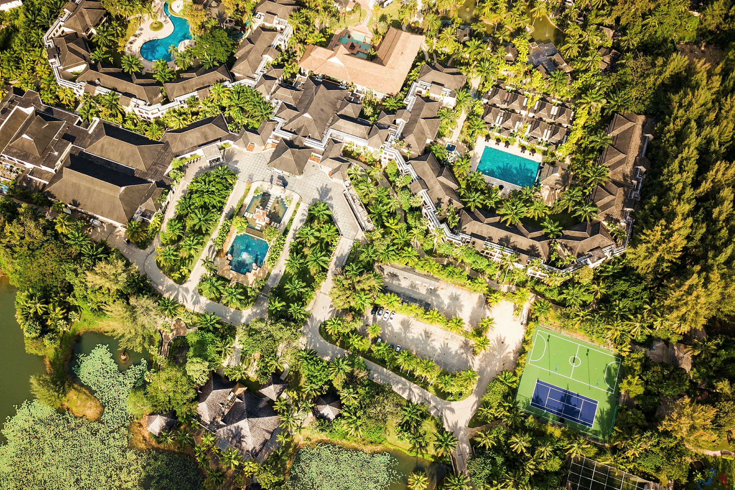 Luftfoto af et resortområde med bygninger, swimmingpools, parkeringsplads, grønne omgivelser og en tennisbane. (AI-genereret tekst)