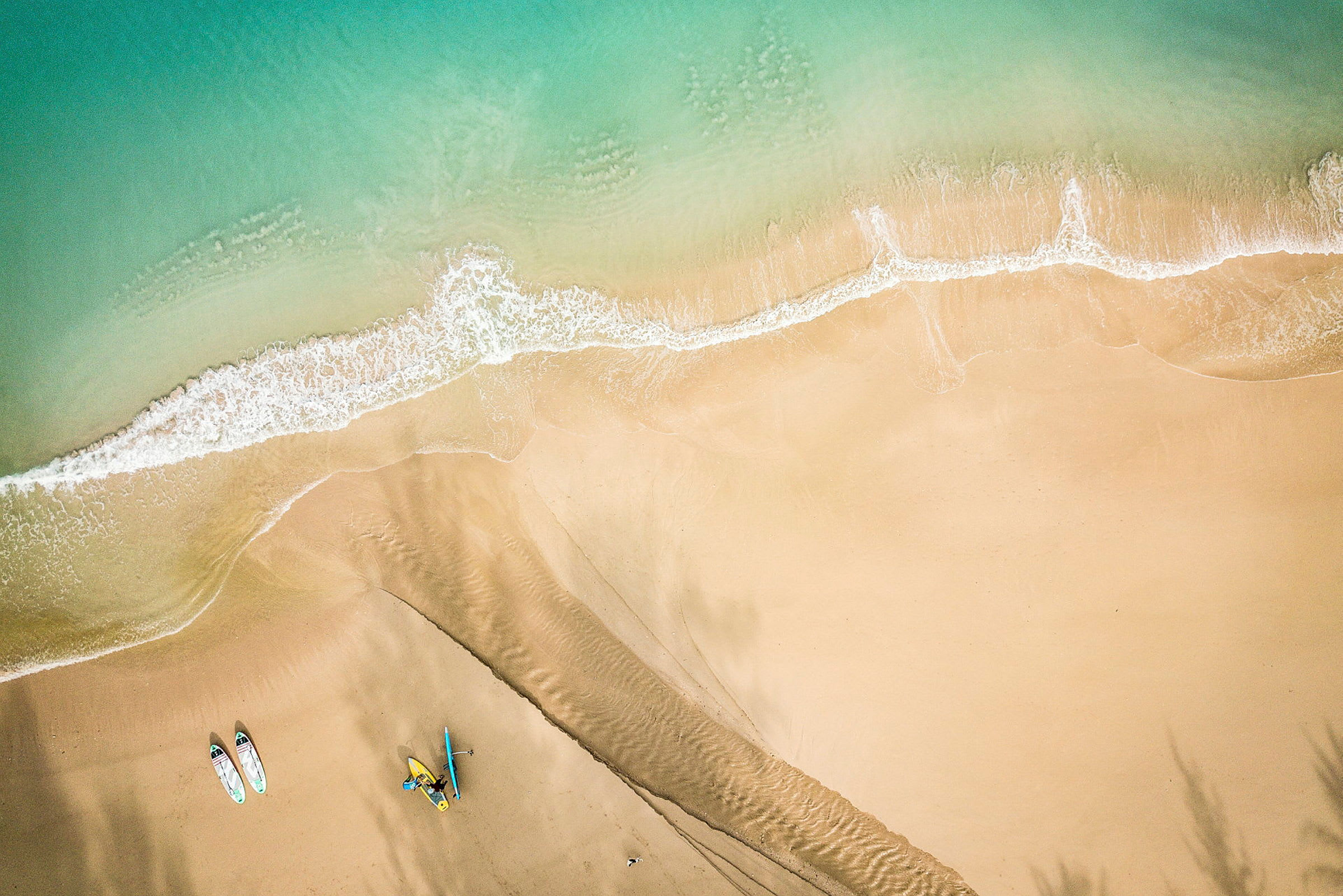 Luftfoto af en sandstrand, hvor bølger møder kysten. To paddleboards og en kajak er synlige på stranden. (AI-genereret tekst)
