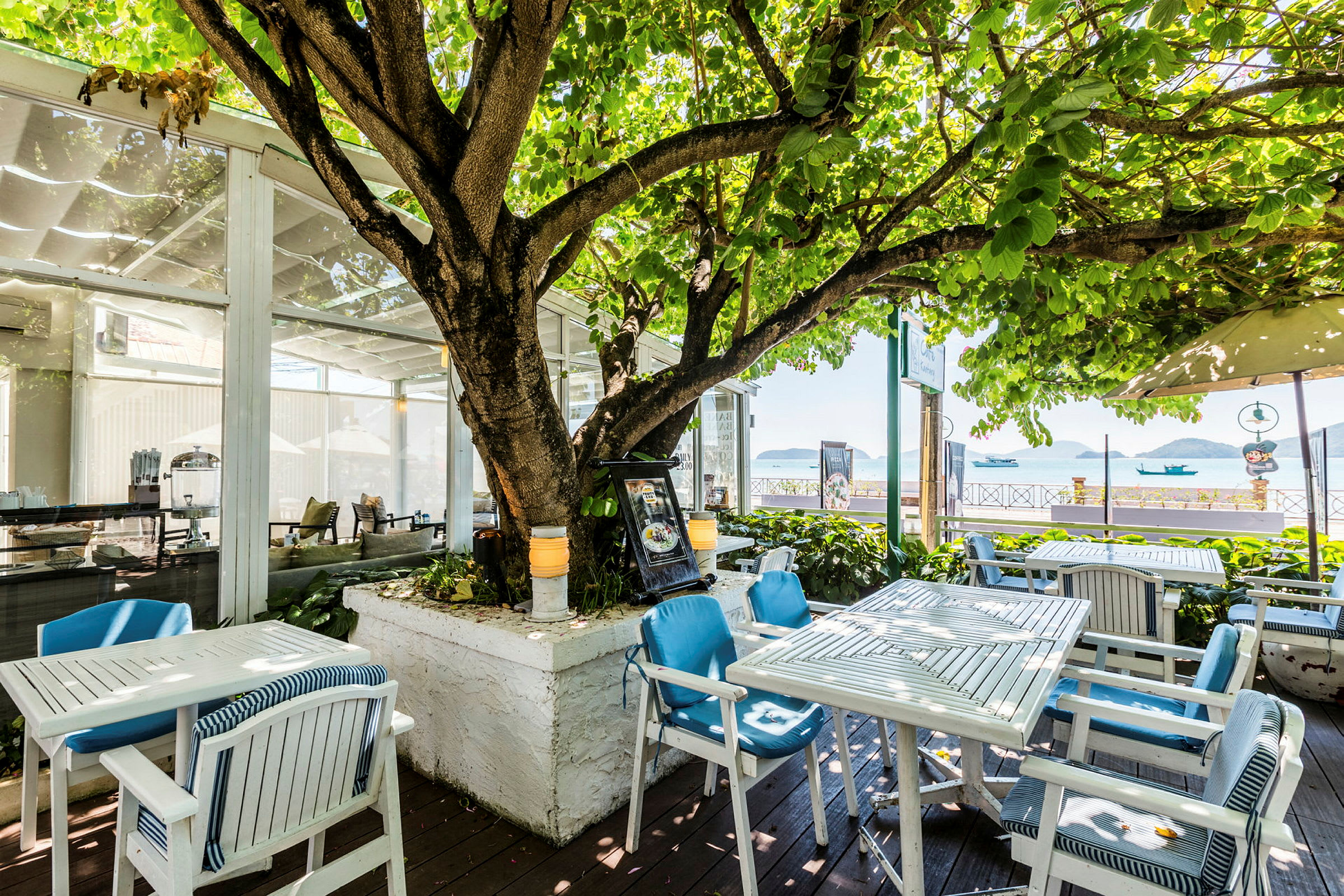Udendørs restaurant med borde og stole under et stort træ ved havet. (AI-genereret tekst)