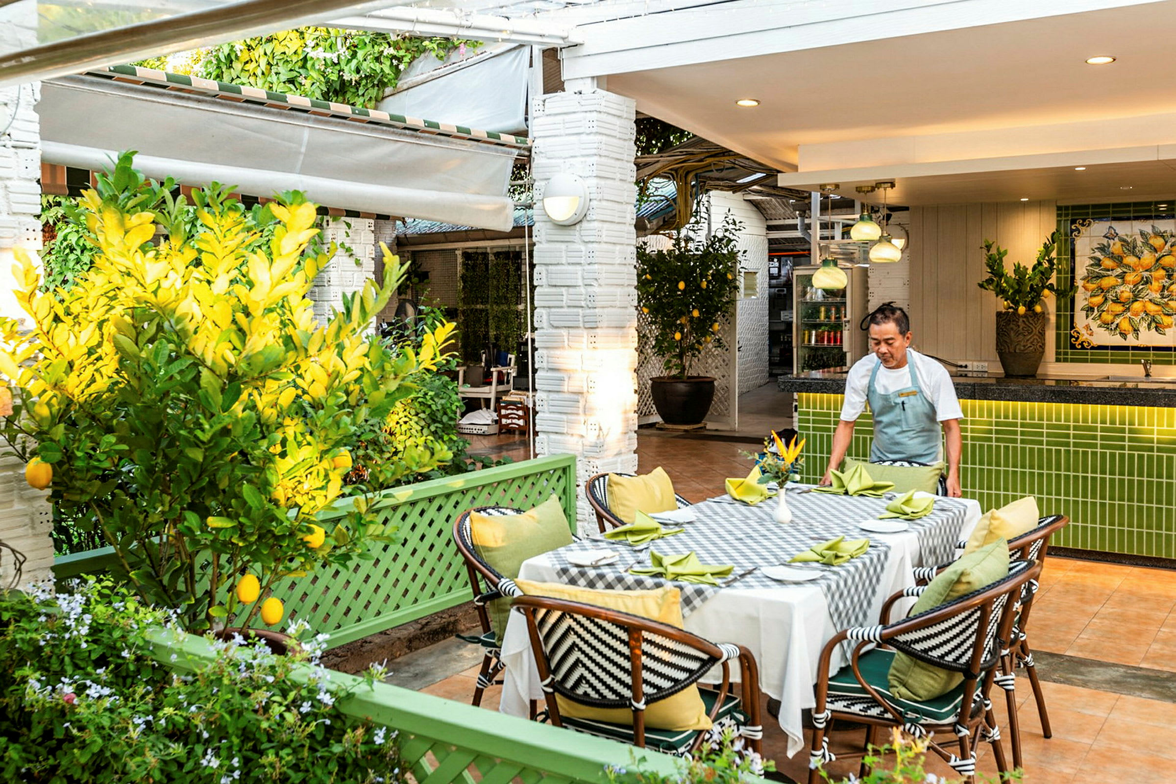En udendørs restaurant med et bord dækket med hvid dug og grønne servietter. En person ordner bordet. Området er omgivet af citrus træer og grønne planter. (AI-genereret tekst)
