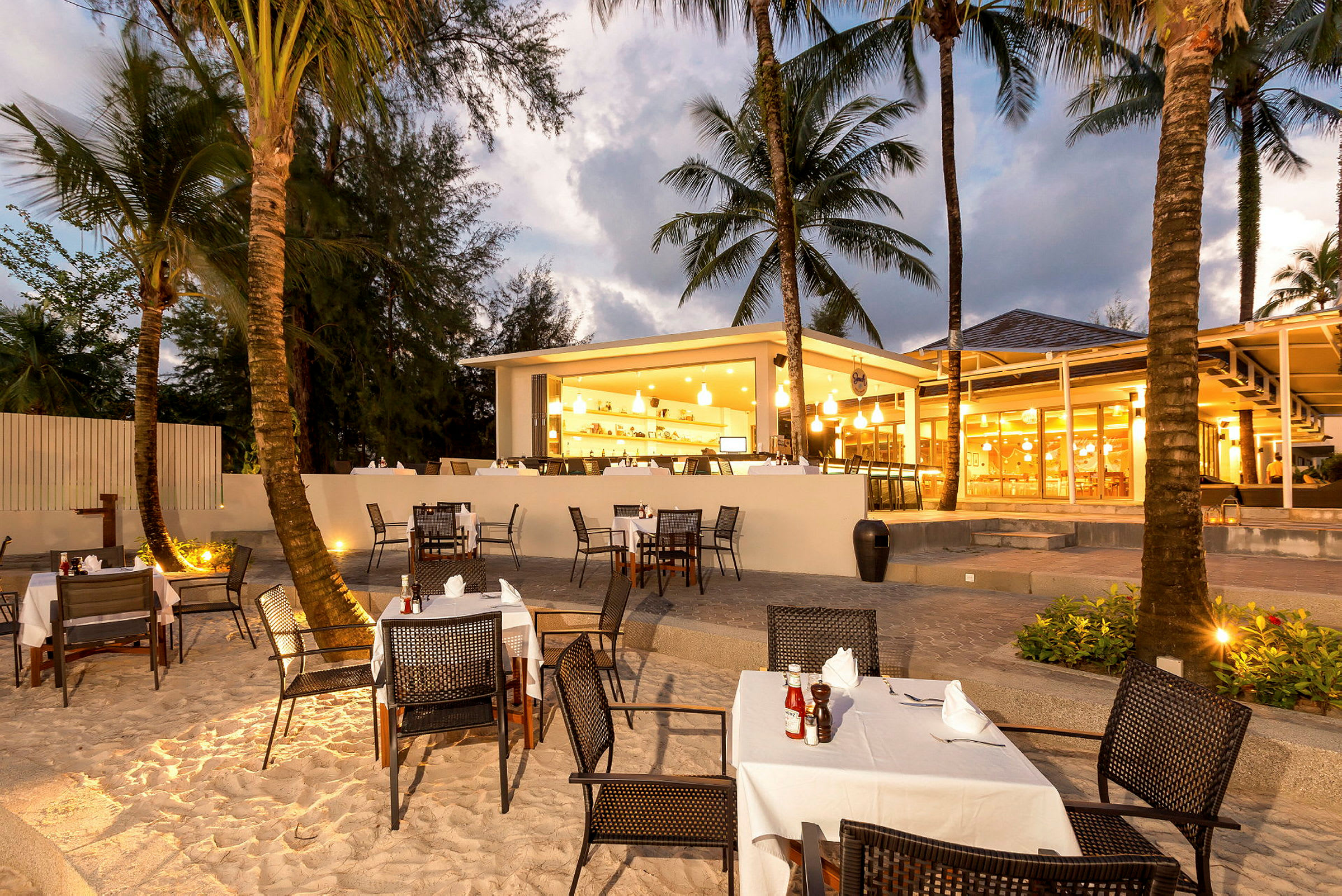Restaurant med udendørs servering ved stranden, omgivet af palmetræer. (AI-genereret tekst)