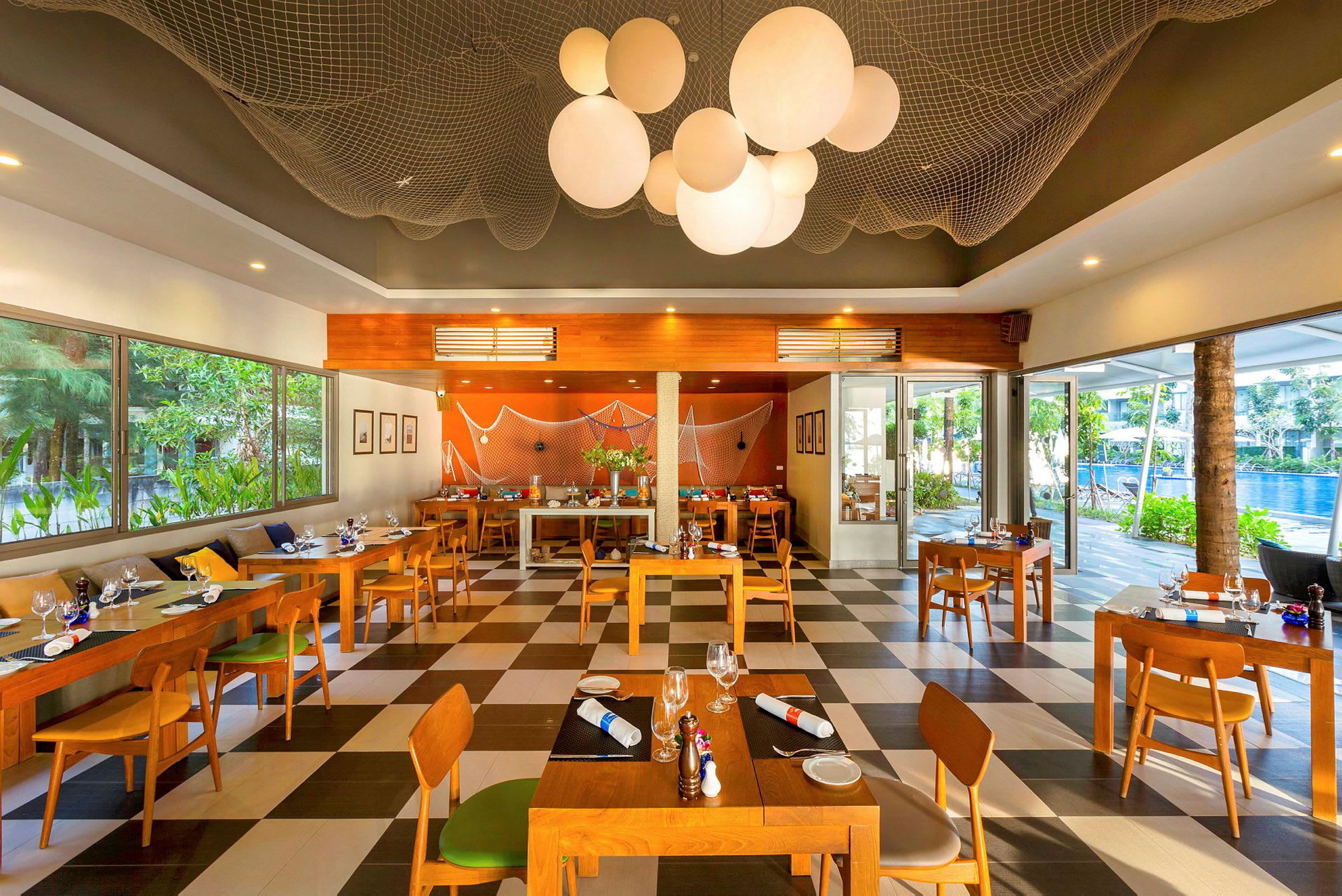 Restaurant med træborde og stole samt store vinduer med udsigt over grønne områder og en pool udenfor. (AI-genereret tekst)