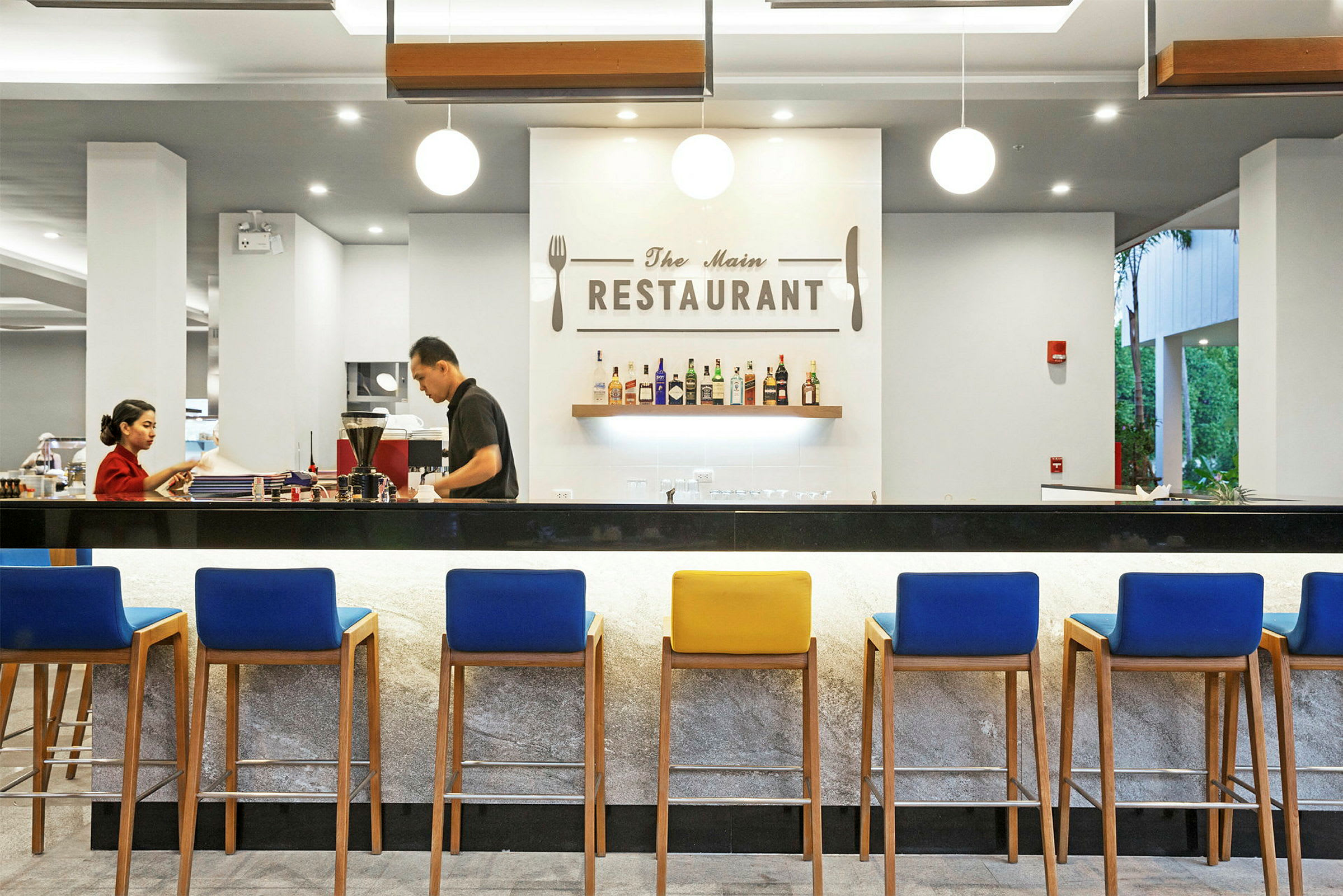 En elegant restaurant med moderne indretning, en bar med farverige stole og en væg med hængte flasker og et skilt med teksten 'The Main Restaurant'. (AI-genereret tekst)