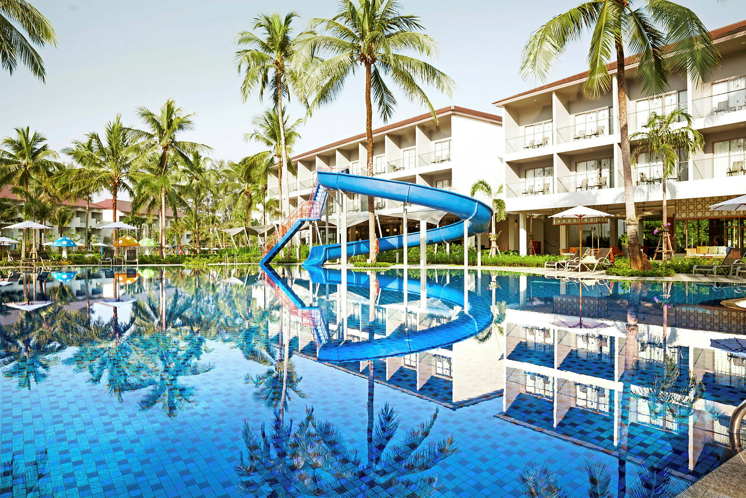 Billede af en pool med en blå vandrutsjebane, omgivet af liggestole og parasoller. Baggrunden viser palmer og en hotelbygnning. (AI-genereret tekst)