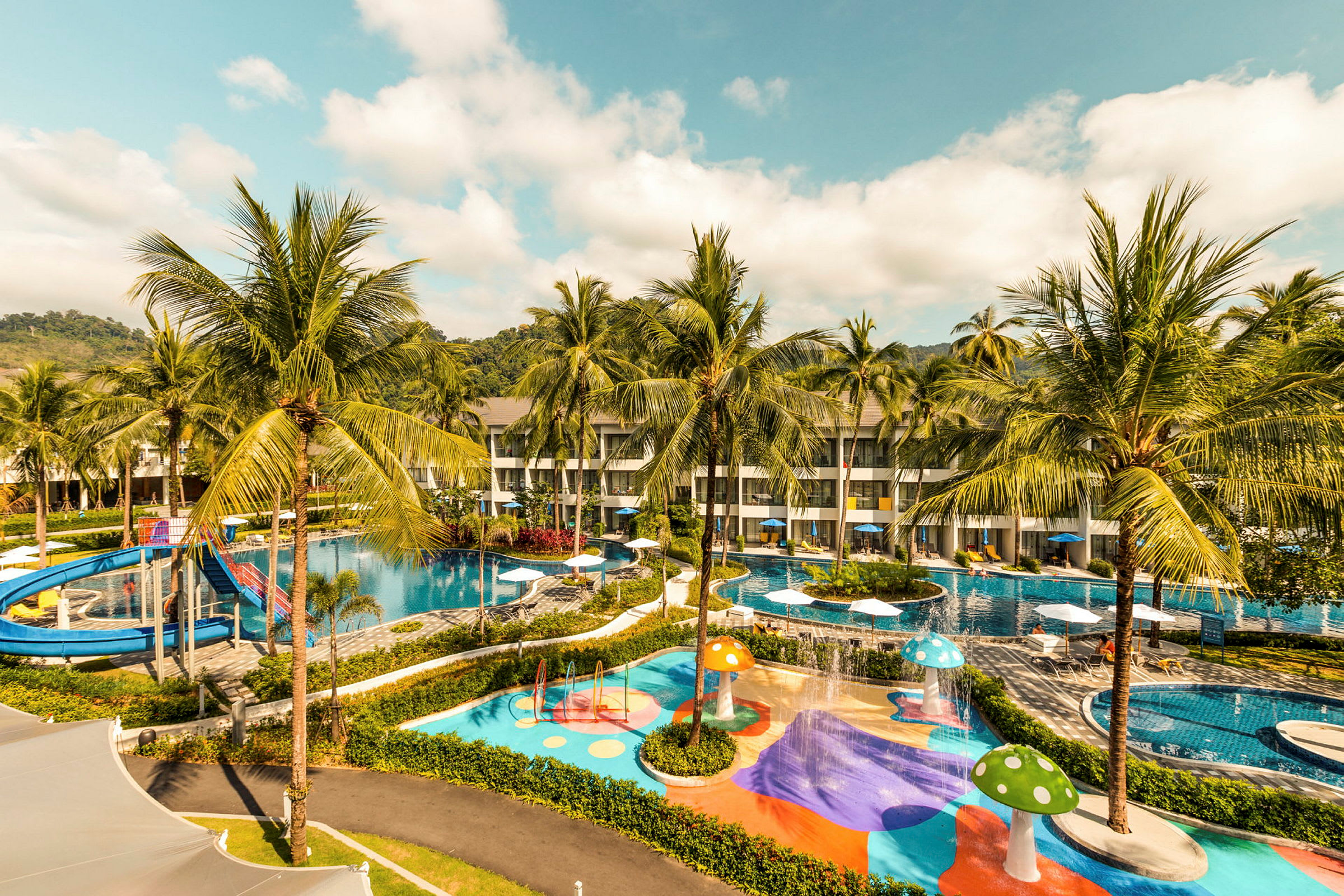 Billede af et hotelområde med swimmingpools, vandrutsjebane og farverige legeområder omgivet af palmer. (AI-genereret tekst)