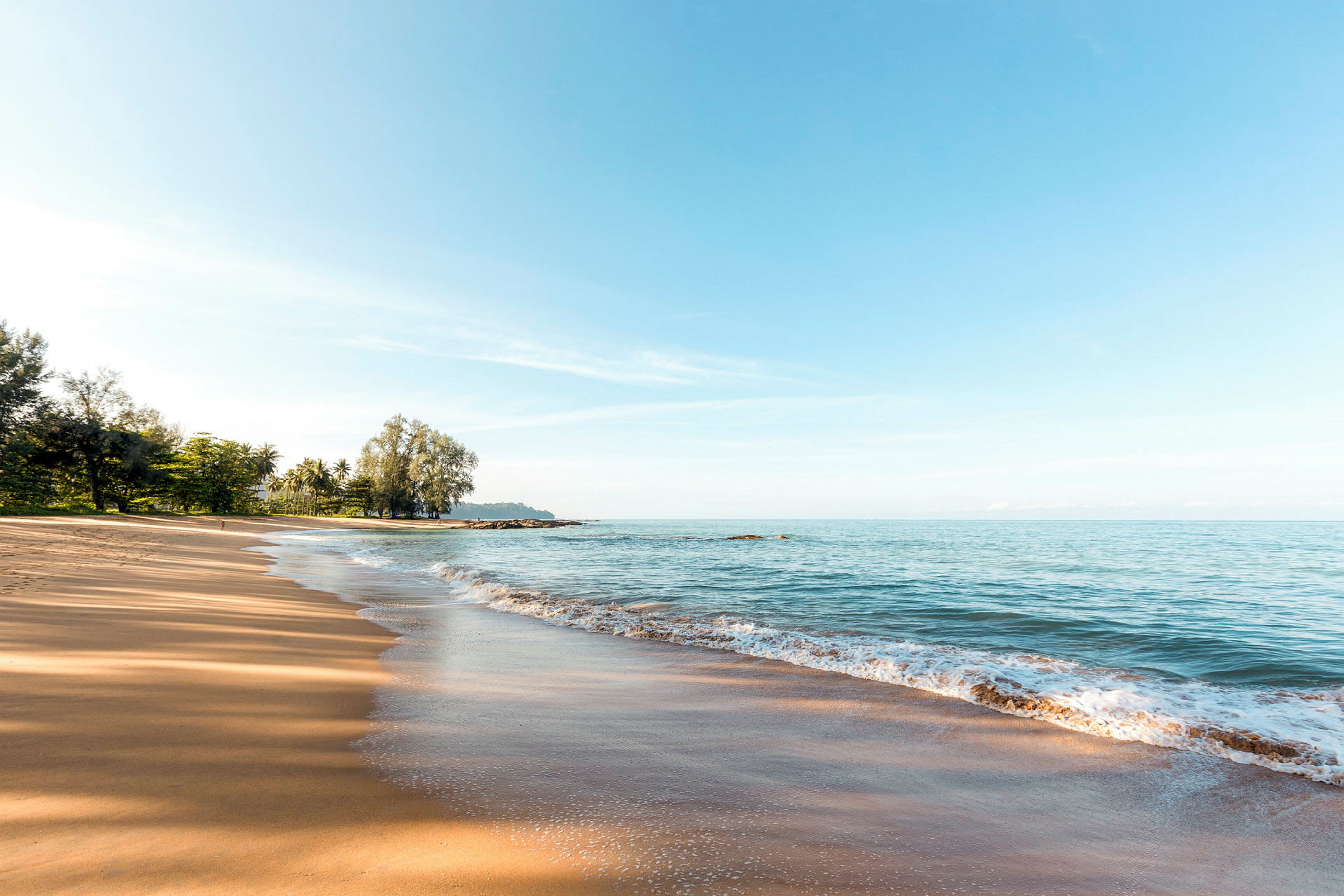 En sandstrand med havet og strandkanten i forgrunden, træer ses i baggrunden. (AI-genereret tekst)