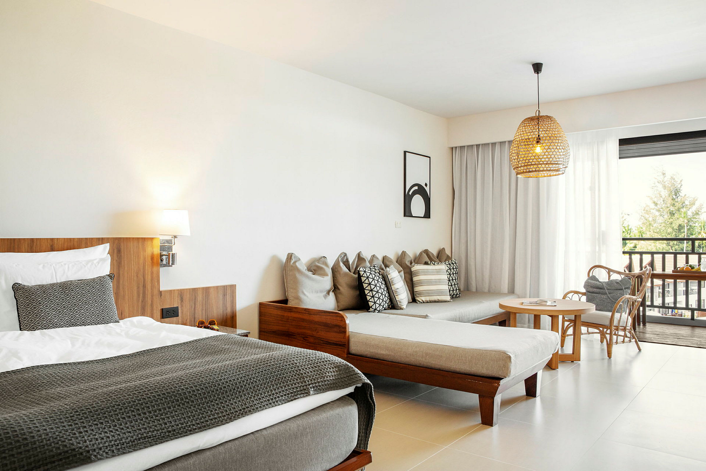 Et hotelværelse med en seng, en sofa med puder, et bord med stole og udsigt udenfor gennem glasdøre. (AI-genereret tekst)
