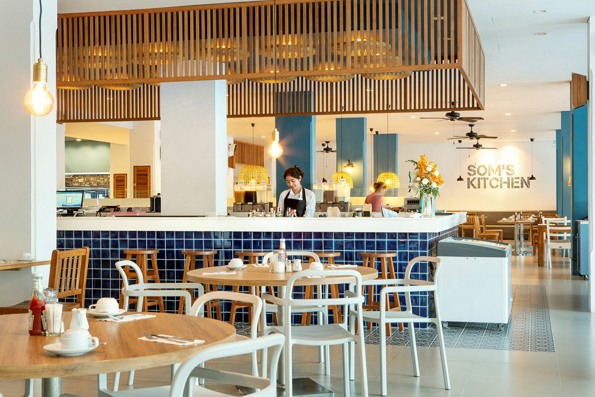 Billede af en restaurant med moderne interiør og blå fliser, hvor træborde og stole er arrangeret rundt omkring. (AI-genereret tekst)