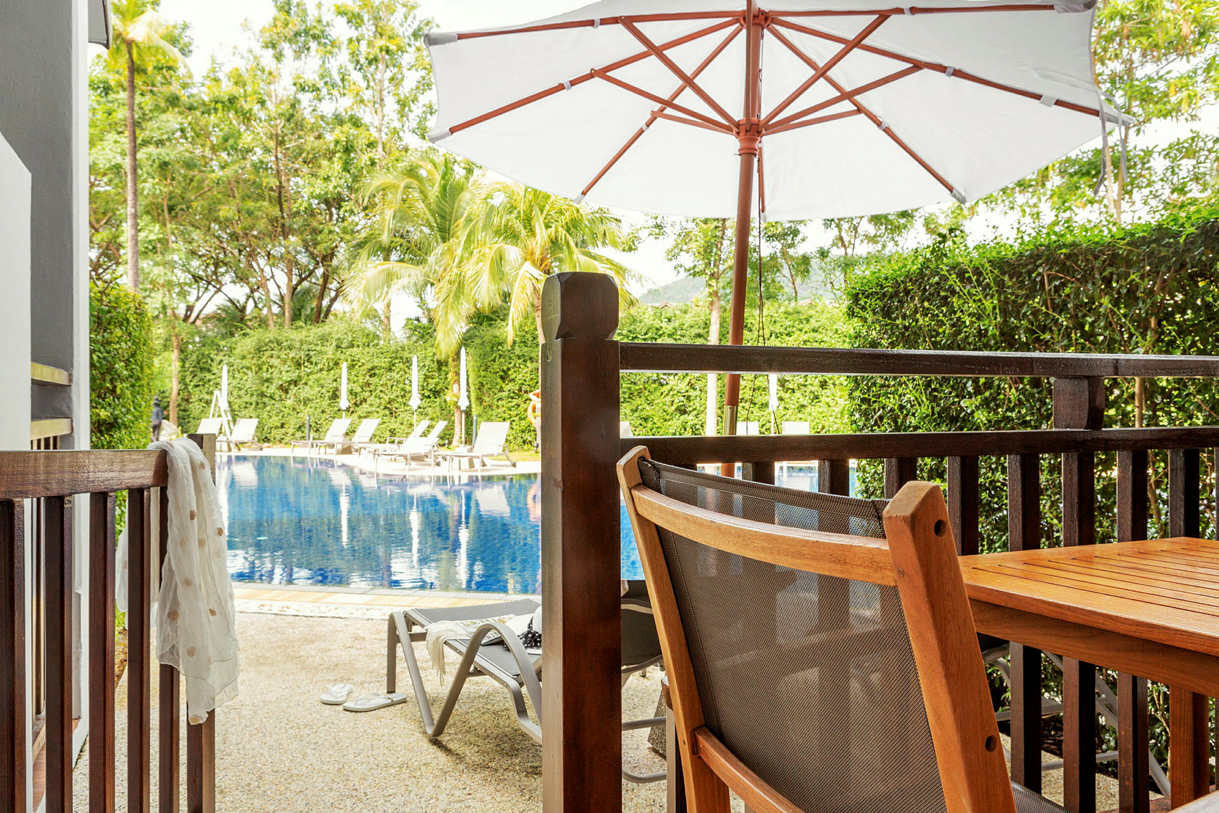 Billedet viser en terrasse med bord og stol af træ tæt på en pool omgivet af liggestole og grønne træer. (AI-genereret tekst)