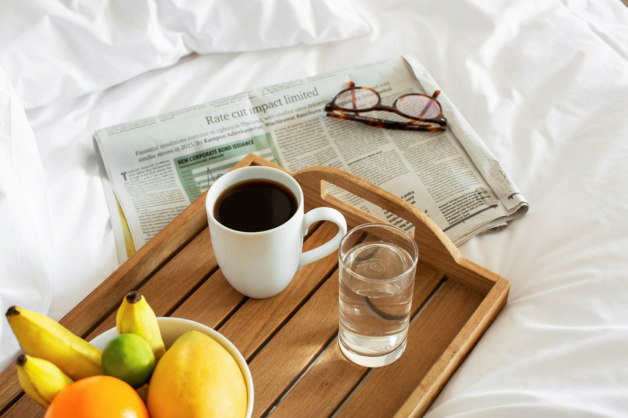 En morgenbakke med en kop kaffe, et glas vand og en skål med frugt på en seng, ved siden af en avis og et par briller. (AI-genereret tekst)