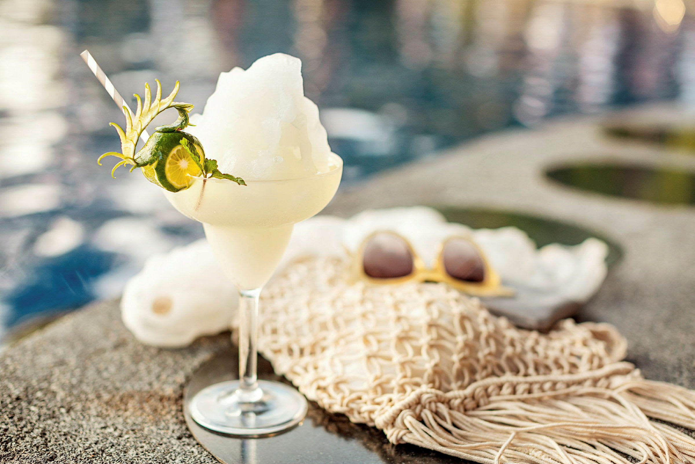 Et glas fyldt med en is-cocktail dekoreret med lime og mynte står på kanten ved siden af en pool. (AI-genereret tekst)