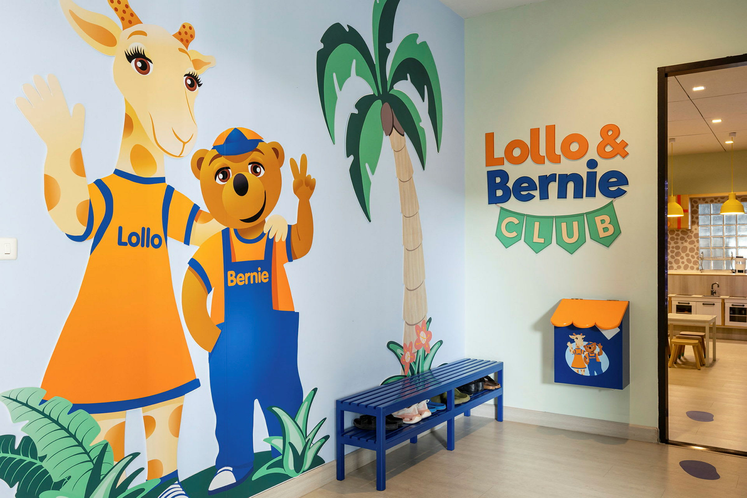 Et farverigt vægmaleri af figurerne Lollo og Bernie står ved en væg i en børneklub. Rummet har også en blå bænk og skilte. (AI-genereret tekst)