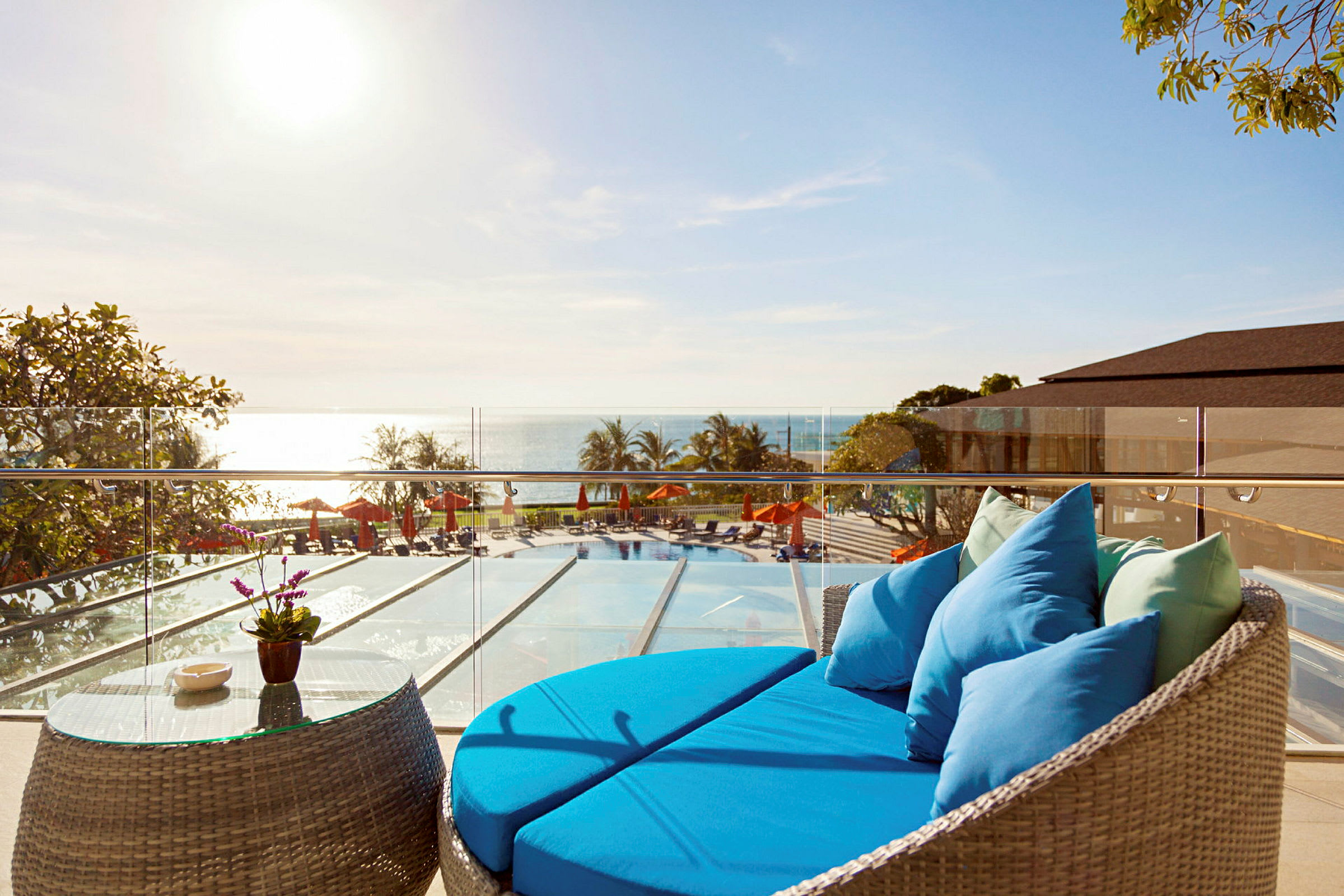 En terrasse med en blå sofa og et glasbord med udsigt over en pool, havet og liggestole under parasoller. (AI-genereret tekst)