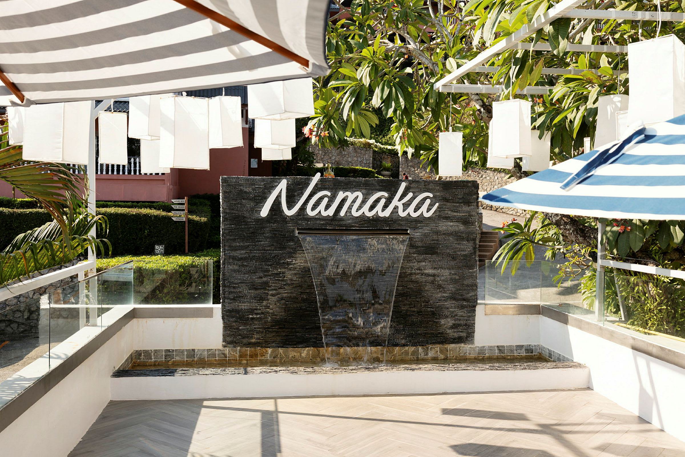 Fontæne med teksten "Namaka" på en sort stenvæg, omgivet af grønne områder og skyggefulde parasoller. (AI-genereret tekst)