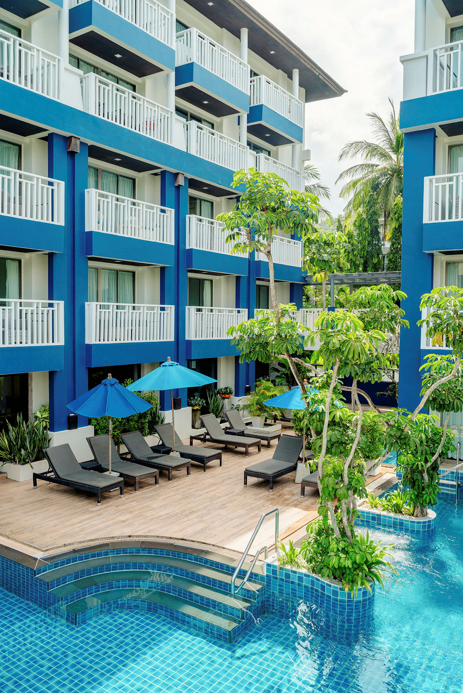 Et hotel med blå facade, pool, liggestole og parasoller omgivet af grønne omgivelser. (AI-genereret tekst)