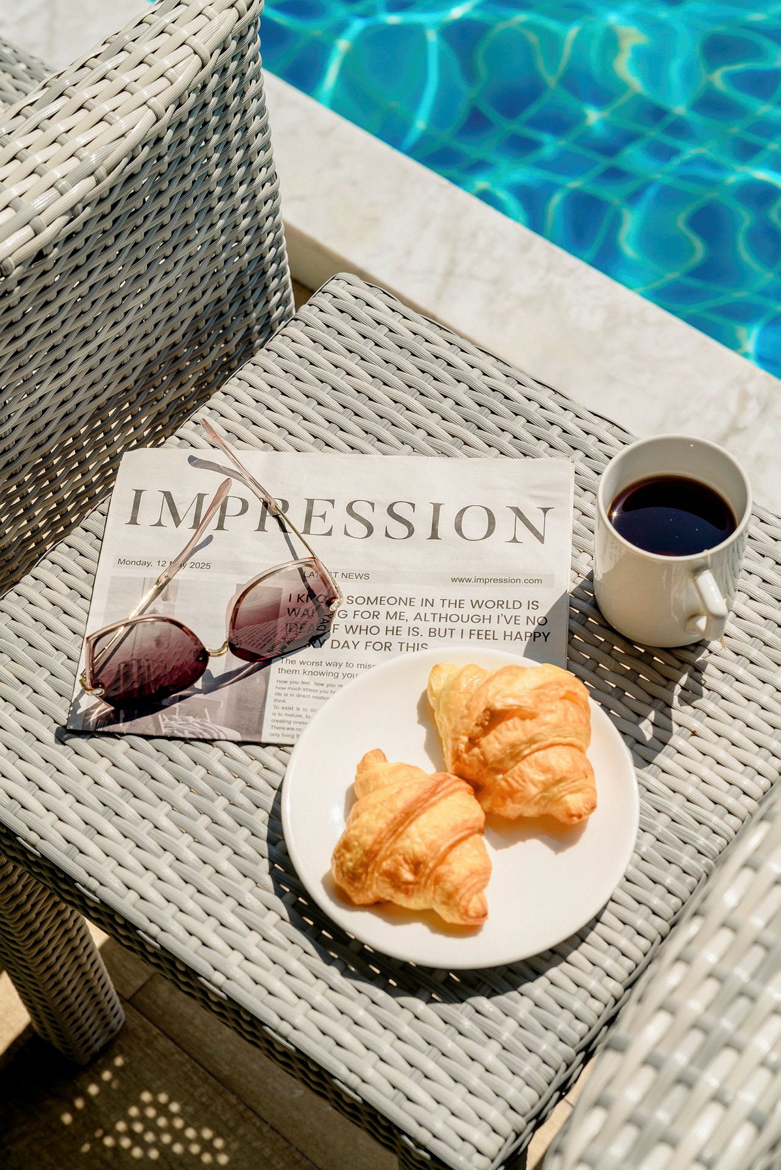 En avis og solbriller på en bordoverflade ved siden af en tallerken med to croissanter og en kop kaffe, med en pool i baggrunden. (AI-genereret tekst)