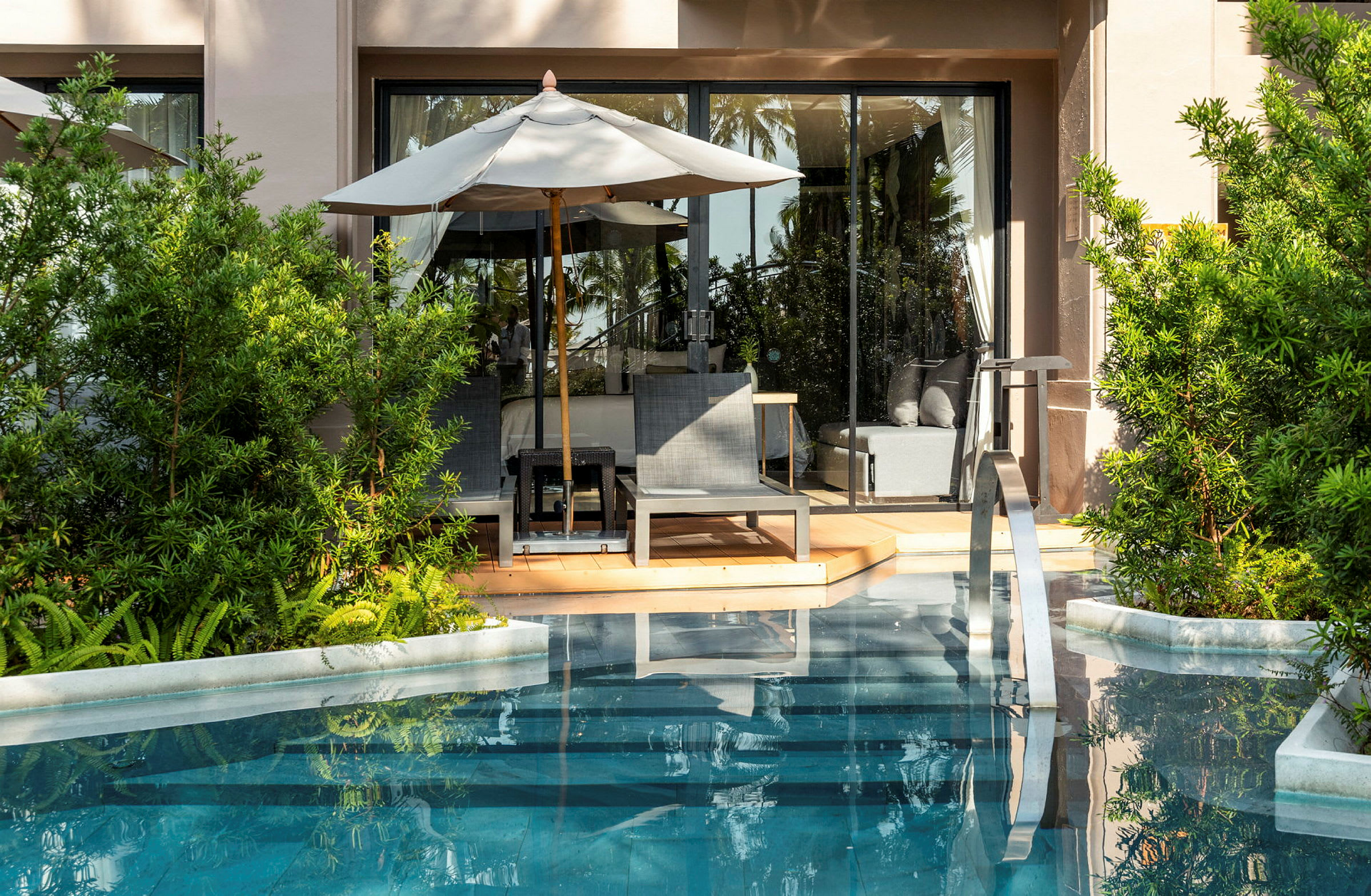 En terrasse med liggestole og parasol ved en pool, omgivet af grønne planter. (AI-genereret tekst)