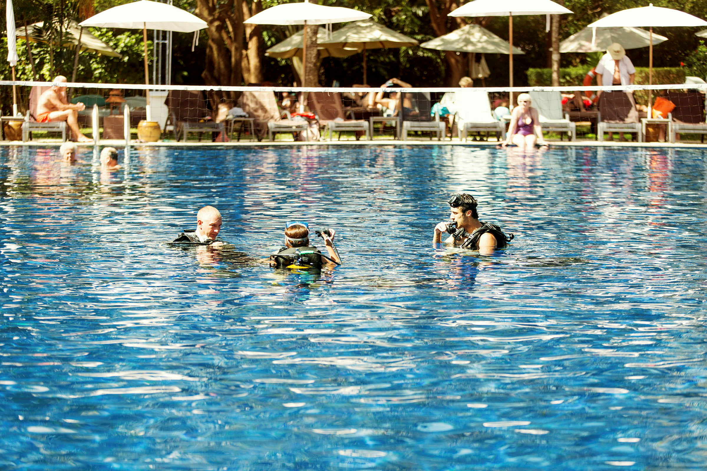 Personer i dykkertøj i en pool med liggestole og parasoller i baggrunden. (AI-genereret tekst)