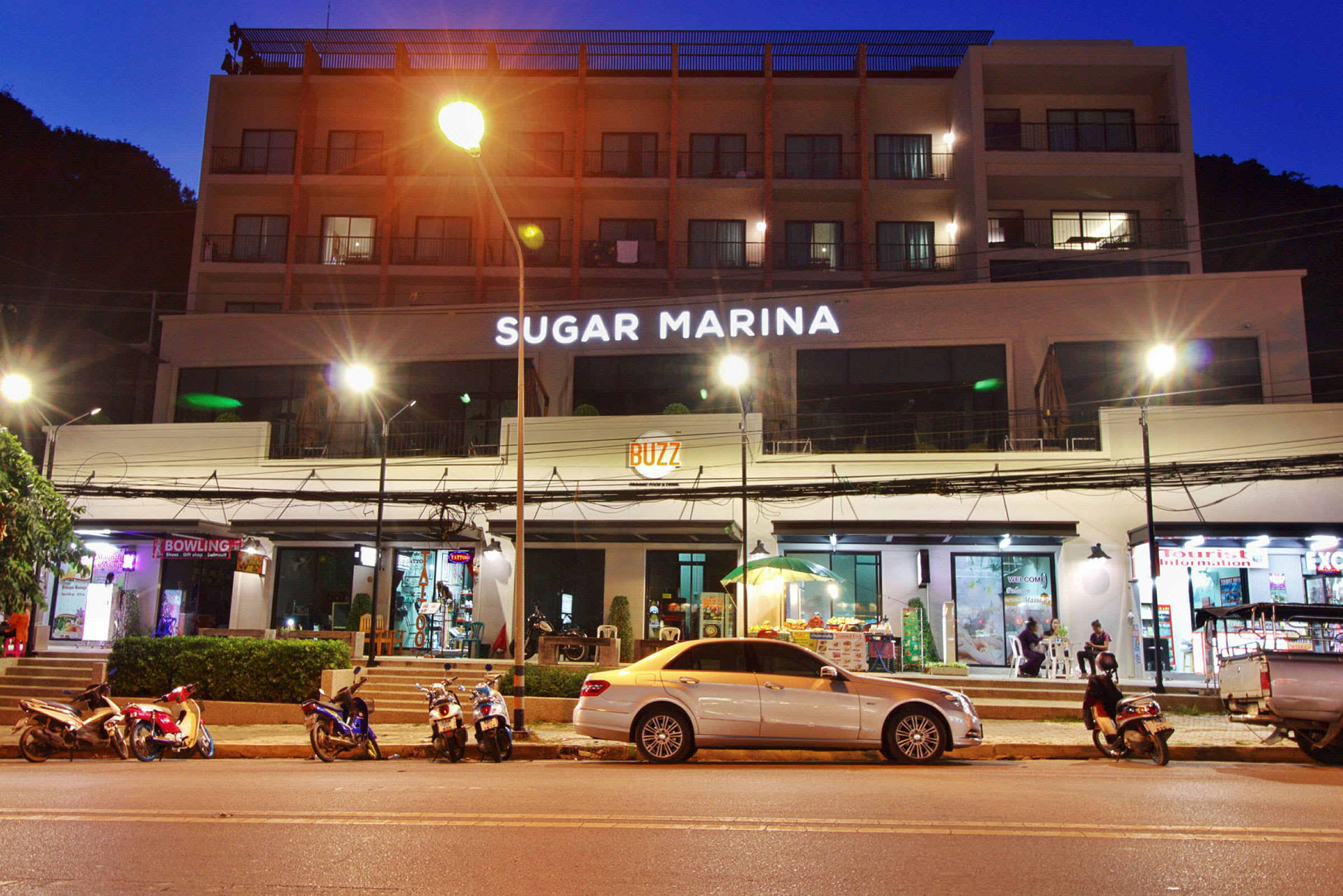 Bygning med hotelnavnet Sugar Marina belyst på facaden om natten, med motorcykler og biler parkeret ved gaden. (AI-genereret tekst)