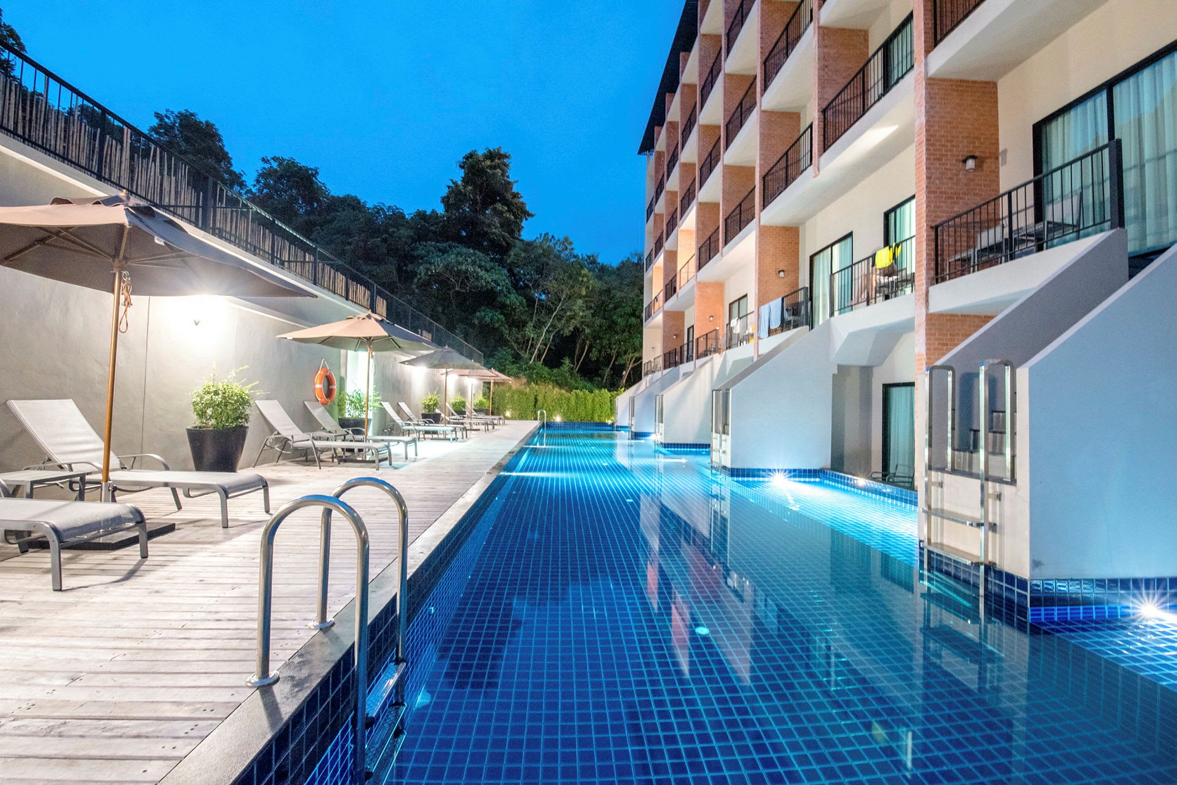 En lang swimmingpool ved et hotel med liggestole og parasoller langs den ene side. (AI-genereret tekst)