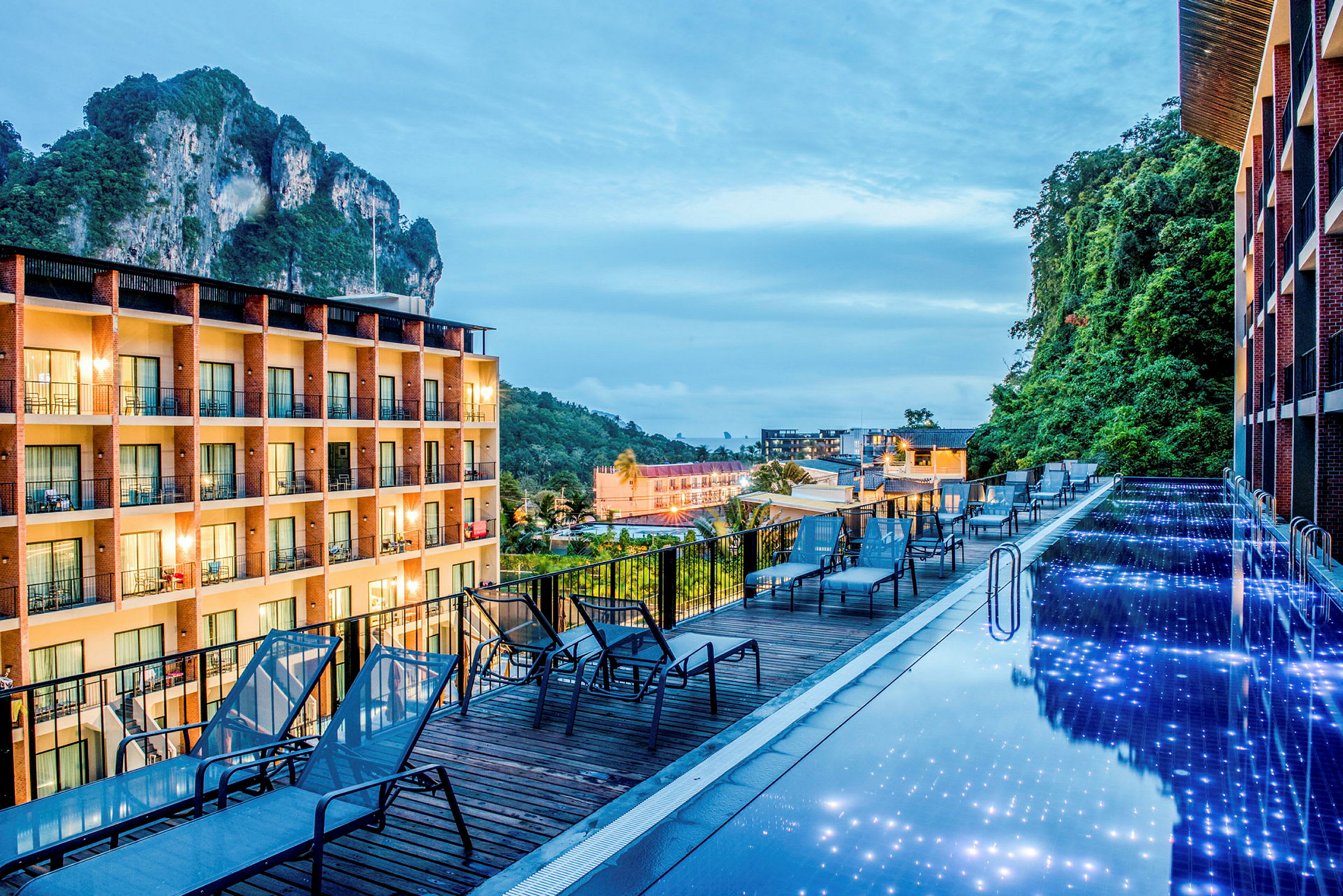 Billede af en pool med lys, der reflekteres i overfladen, omgivet af liggestole og en treetagers bygning i baggrunden. (AI-genereret tekst)