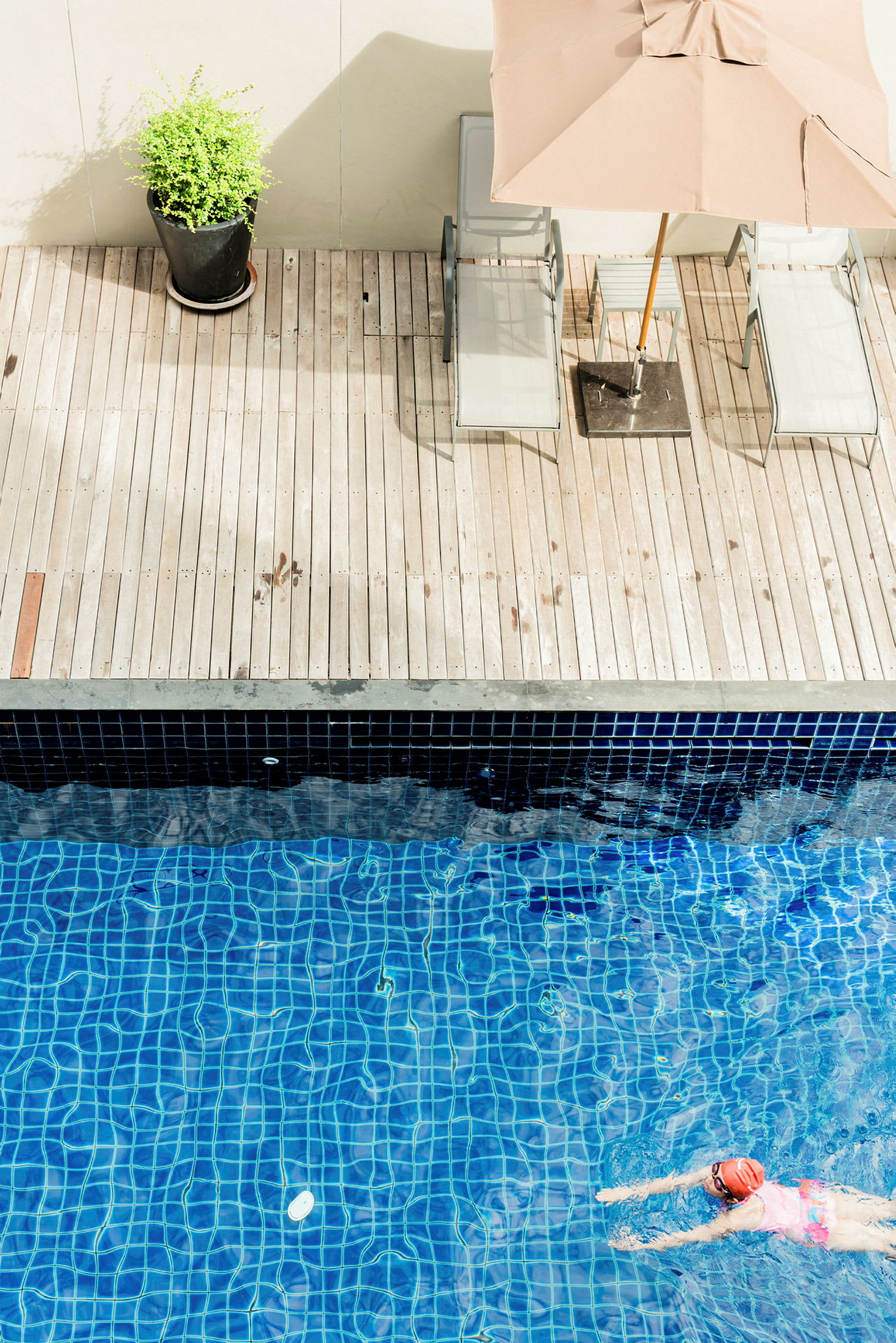 En person svømmer i en pool med blå fliser. Ved siden af poolen er der en træterrasse med liggestole, en parasol og en potteplante. (AI-genereret tekst)