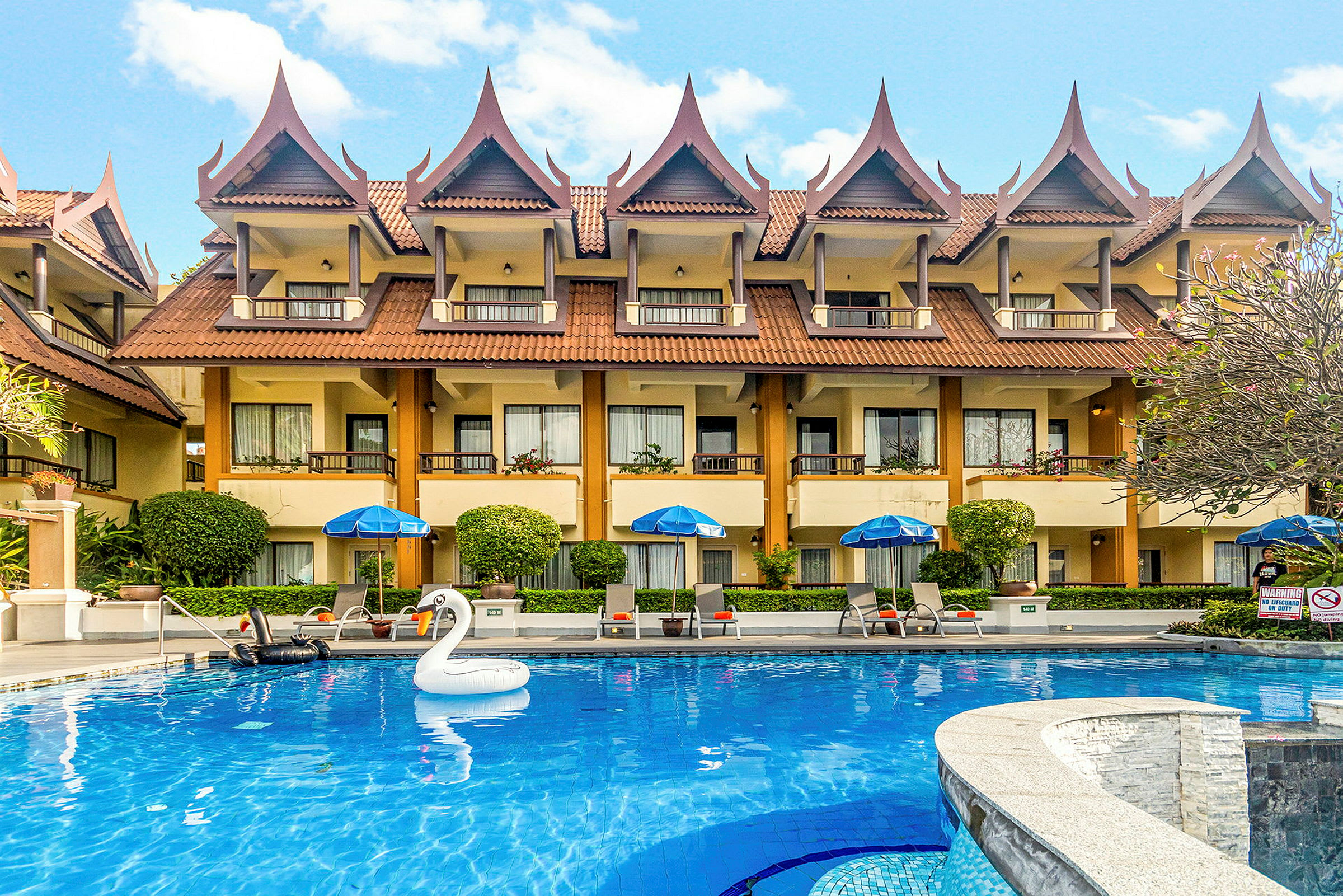 Et hotel med thailandske tagdetaljer og en blå pool foran, inklusive en oppustelig svane. (AI-genereret tekst)