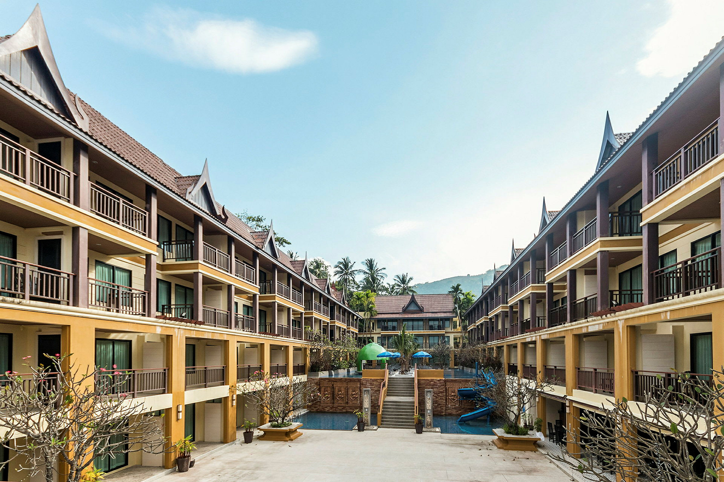 Billede af et hotel med flere etager og balkoner omkring et centralt poolområde med træer og parasoller. (AI-genereret tekst)