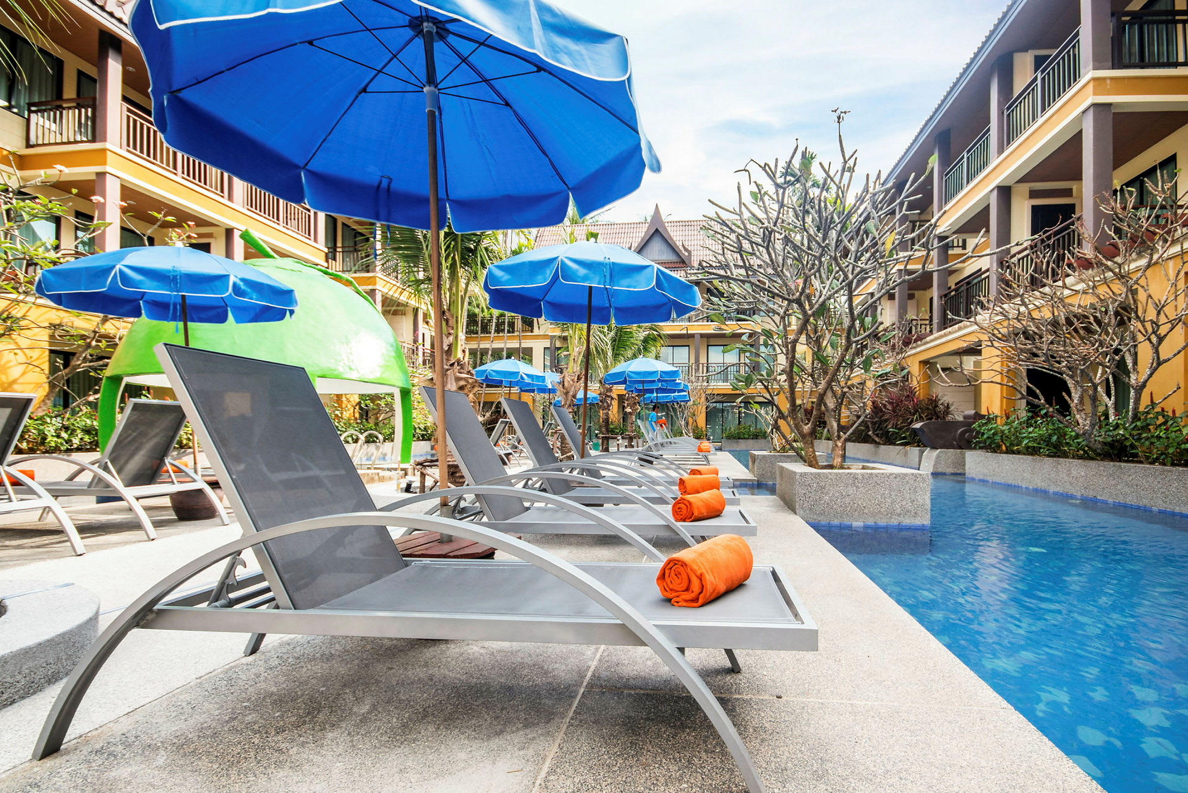 Liggestole med orange håndklæder under blå parasoller ved en pool omgivet af hotelbygninger. (AI-genereret tekst)