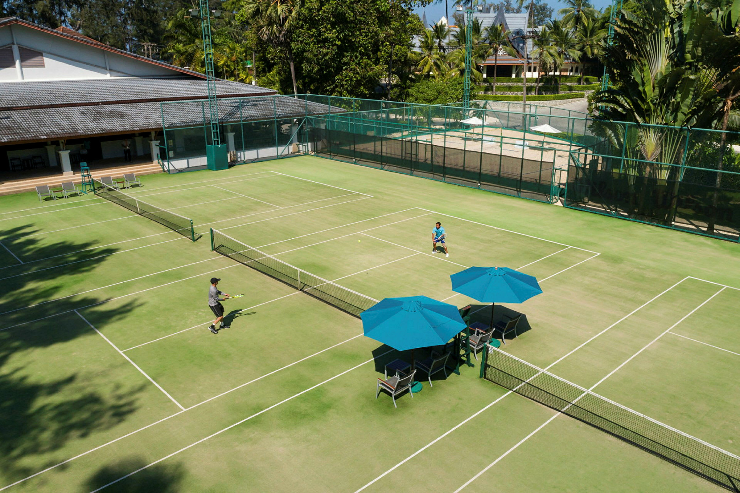 En grøn tennisbane med to personer, der spiller tennis. Der er blå parasoller og stole ved siden af banen. (AI-genereret tekst)