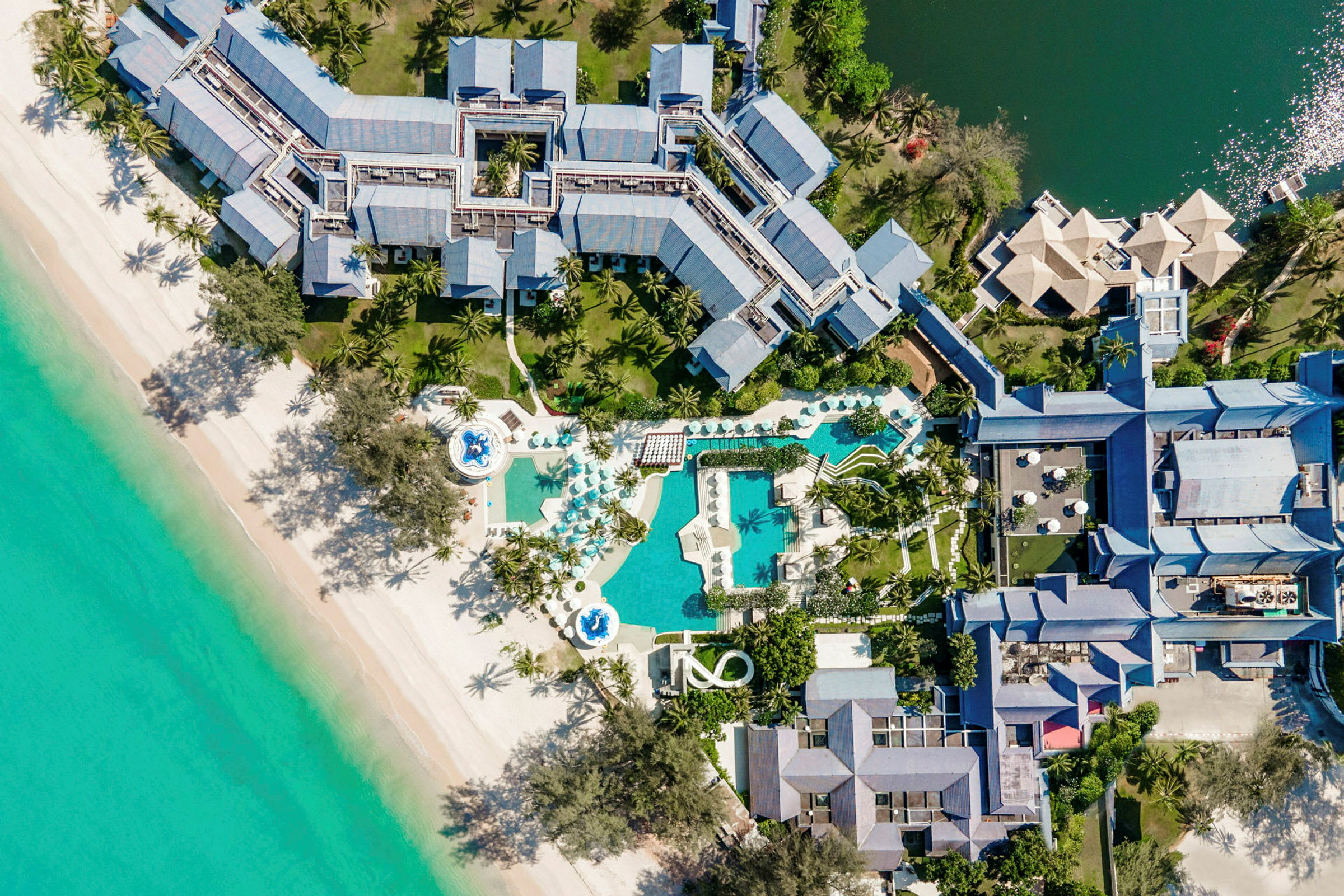 Luftfoto af et feriested med poolområder og palmer ved en strand. (AI-genereret tekst)