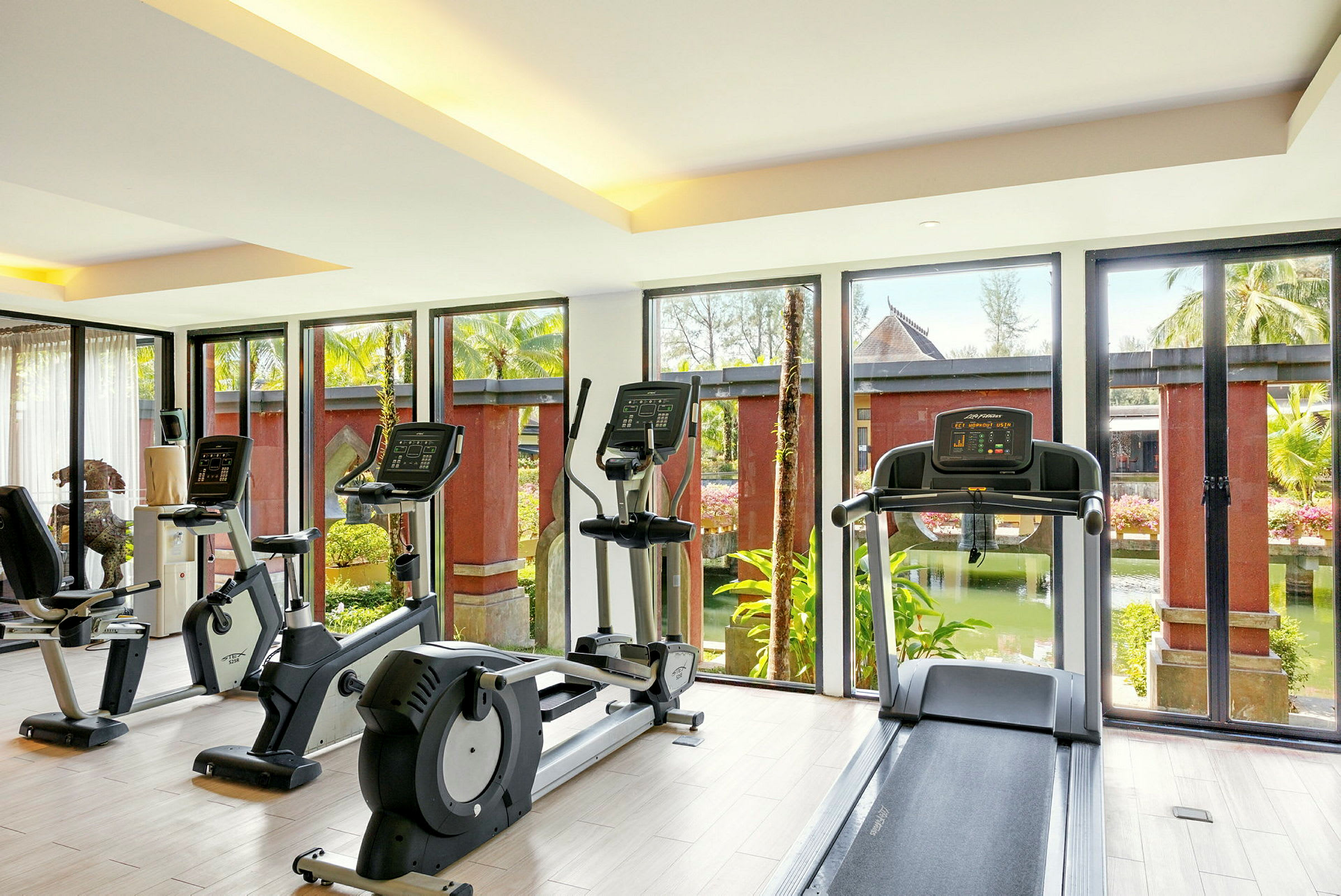 Et fitnesscenter med træningsmaskiner foran store vinduer ud mod en have. (AI-genereret tekst)