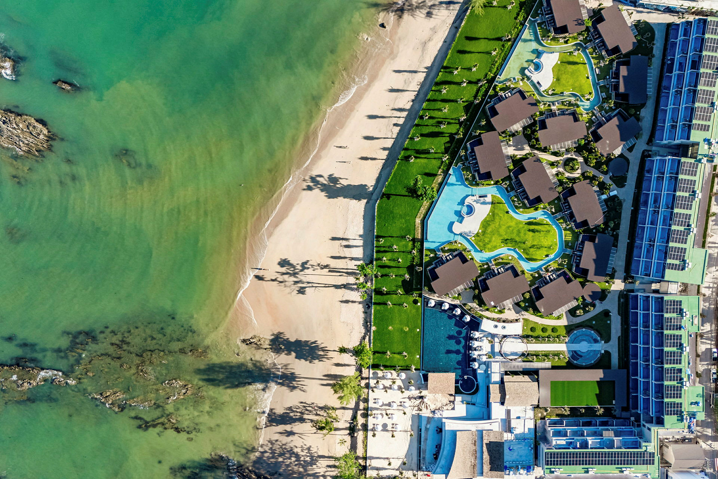 Luftfoto af en strand og et hotelområde med pools og bygninger. (AI-genereret tekst)