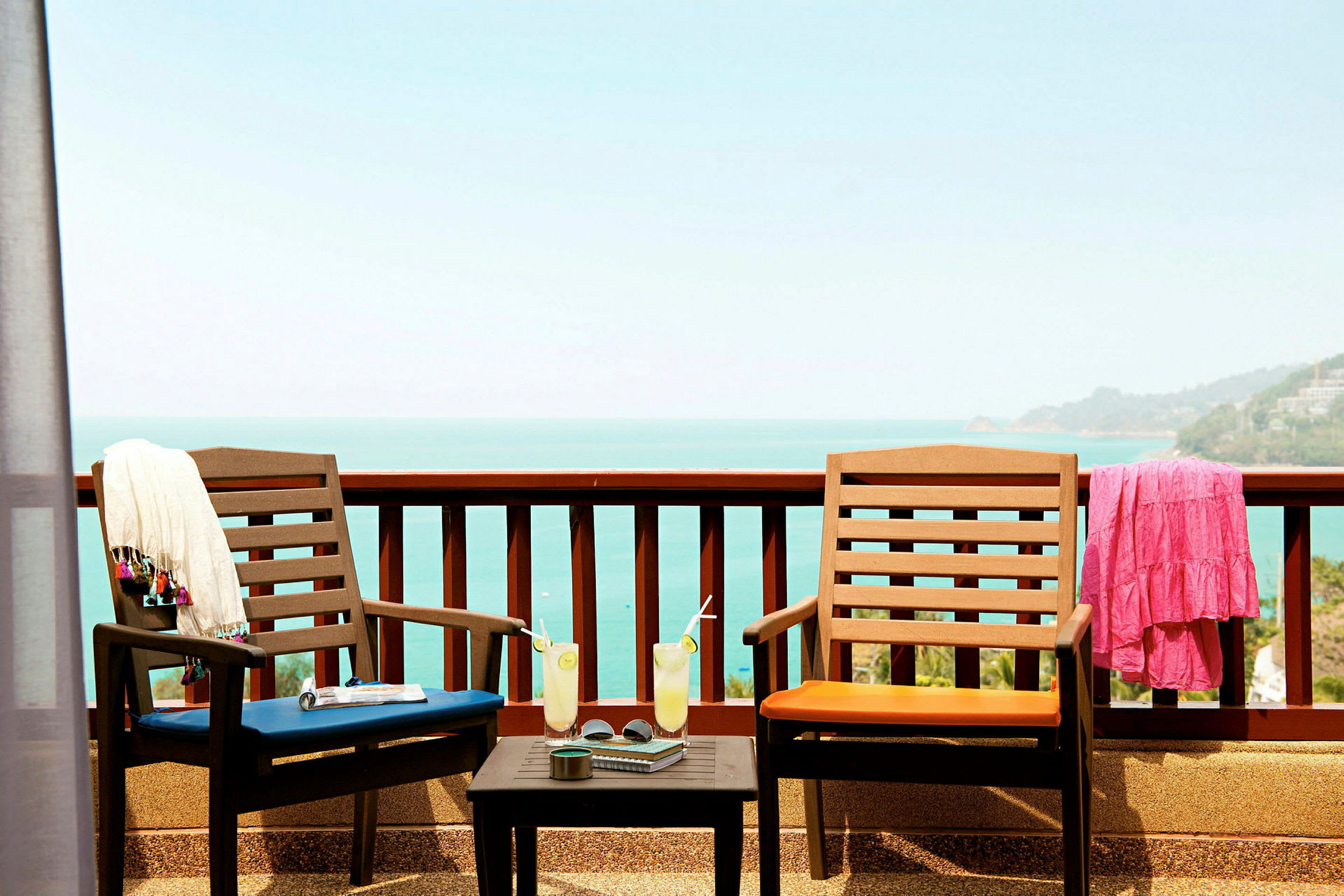 To udendørsstole med hynder står på en terrasse med udsigt over havet. På bordet mellem stolene står glas med drikke og en kop. Et tørklæde og en kjole hænger på stoleryggene. (AI-genereret tekst)