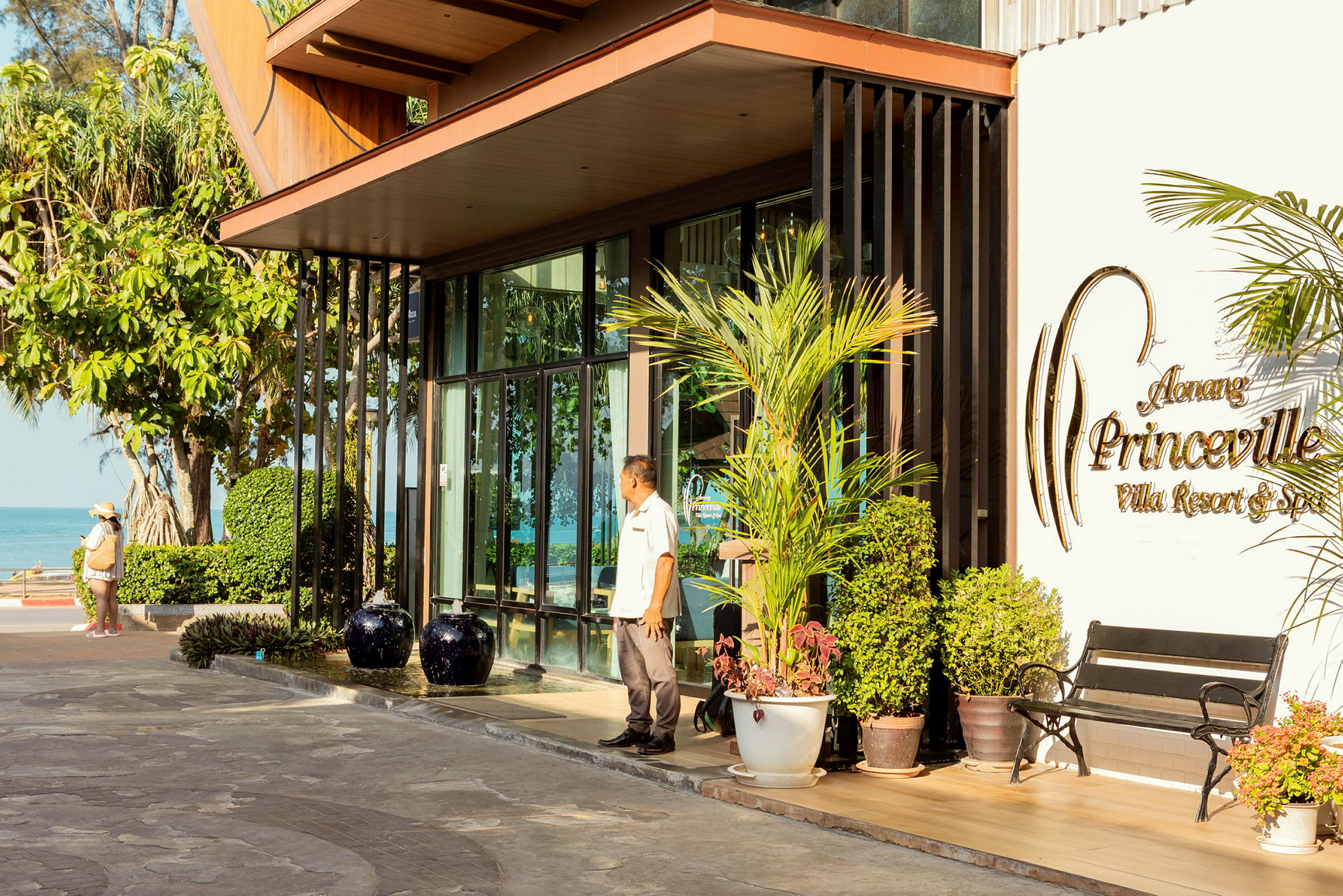 En indgang til en resort med skiltet "Aonang Princeville Villa Resort & Spa". Udendørs er der beplantning og en bænk, med baggrund af hav og grønne områder. (AI-genereret tekst)