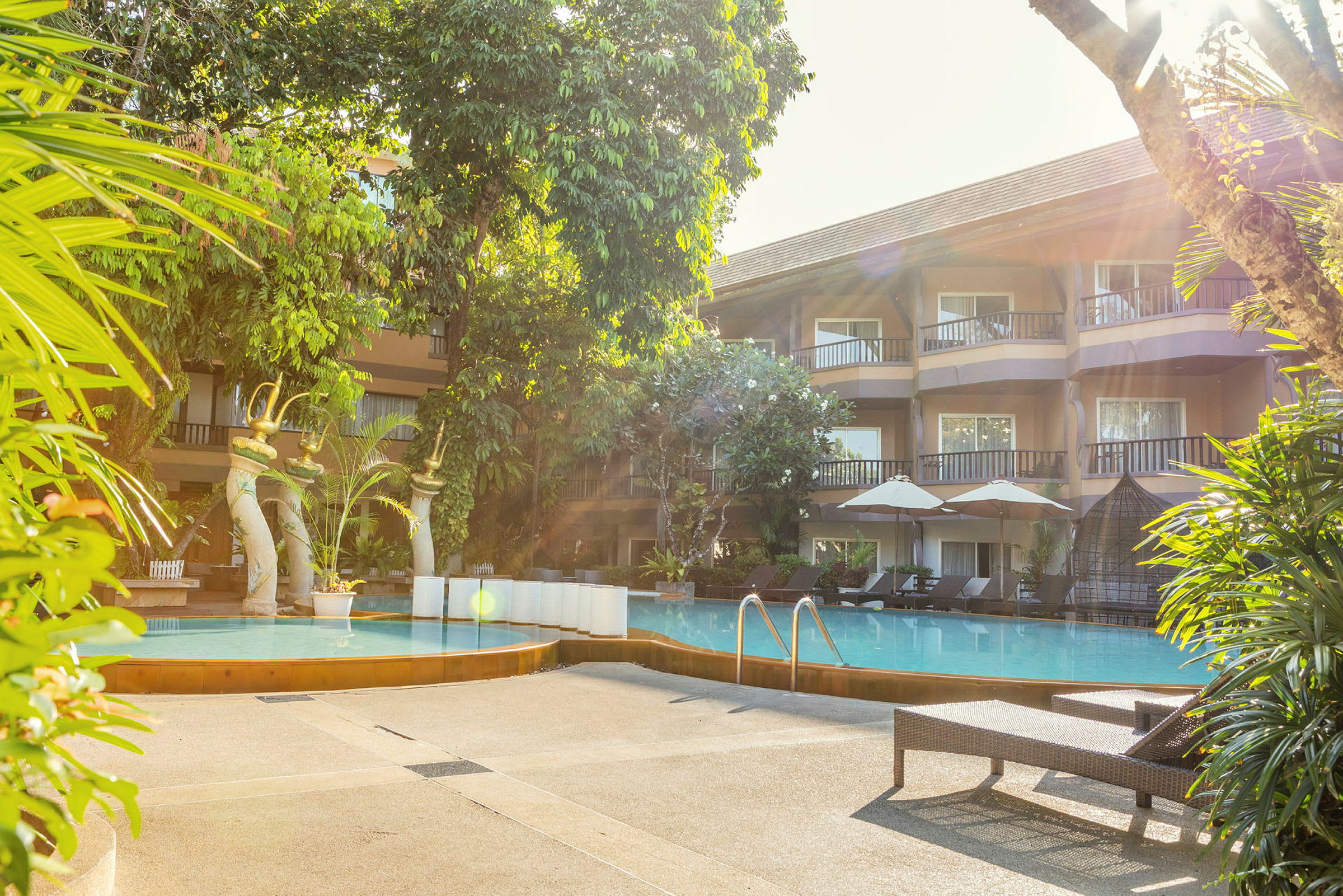 Billede af en hotelpool omgivet af liggestole og parasoller, med frodig vegetation og bygningen i baggrunden. (AI-genereret tekst)