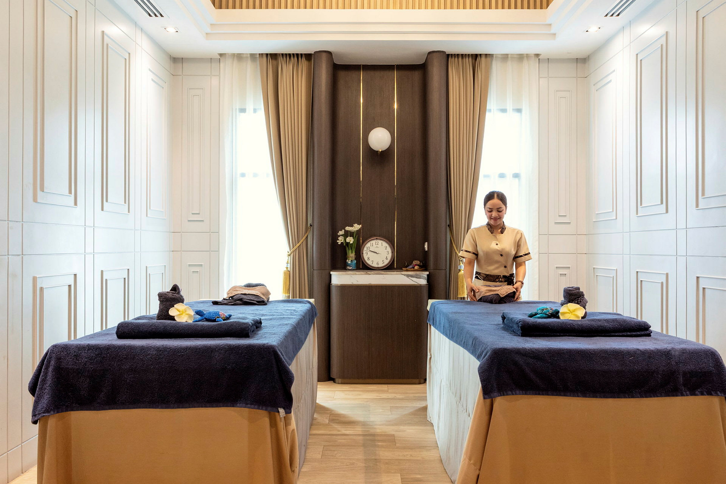 Billede af et spa-rum med to massageborde dækket med mørkeblå håndklæder og en person, der forbereder behandlinger. (AI-genereret tekst)