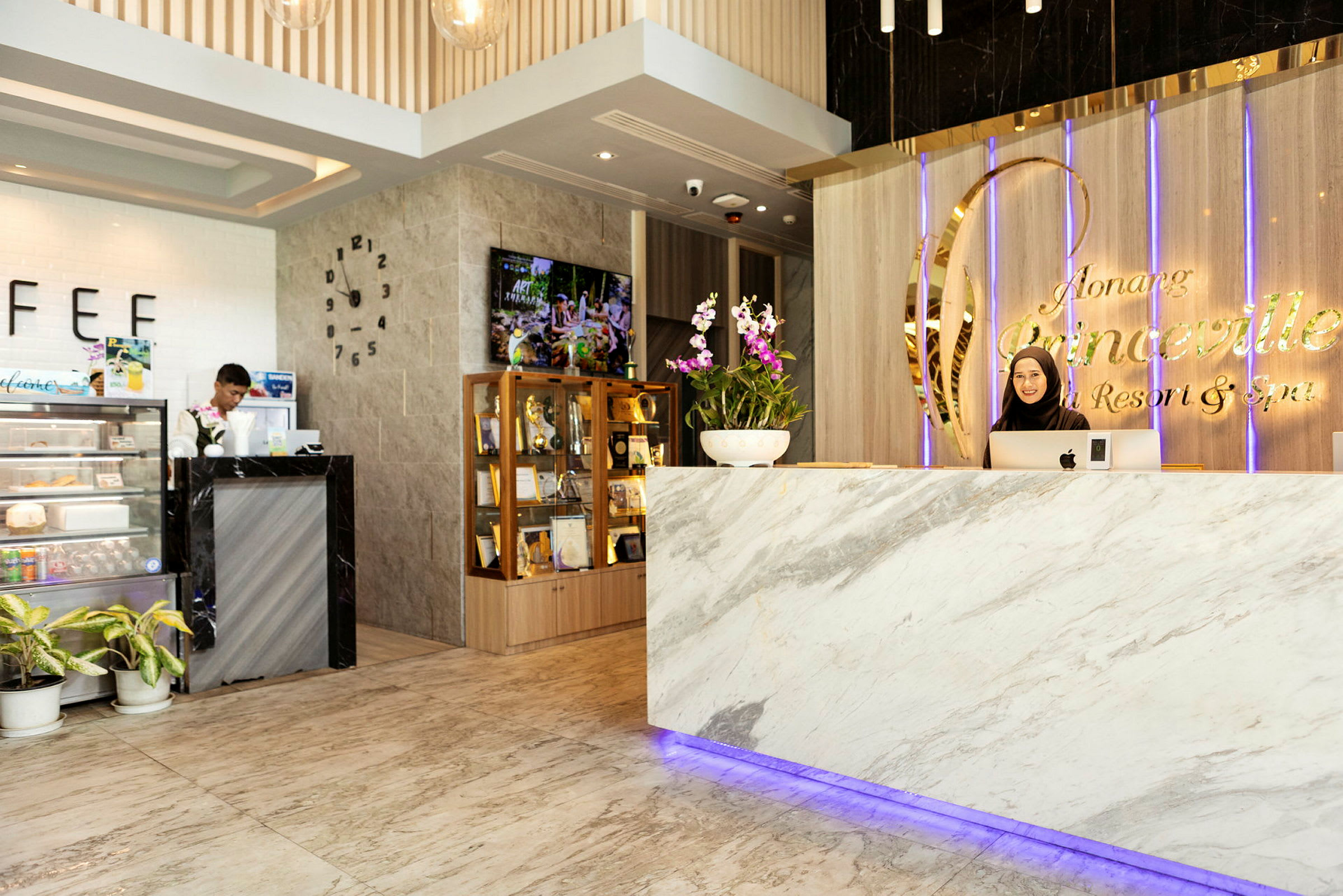 Reception med marmordisk og skilt med teksten "Ao Nang Princeville Resort & Spa". En person står bag disken. Til venstre ses en lille cafédisk og et vitrineskab med priser. (AI-genereret tekst)