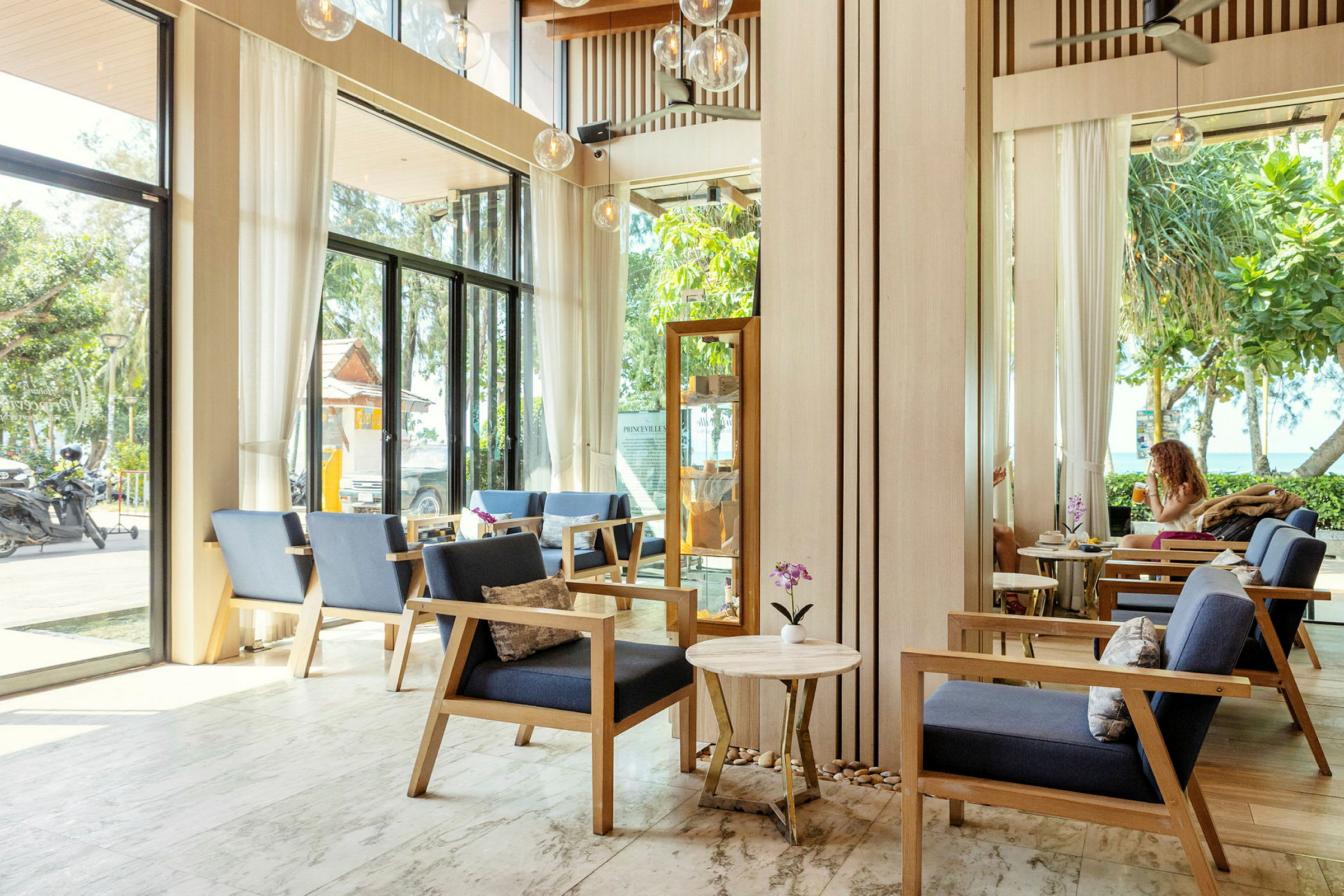 Billede af en lys og moderne lounge indrettet med behagelige træstole, borde og glasvægge med udsigt til grønne områder og en vej udenfor. (AI-genereret tekst)