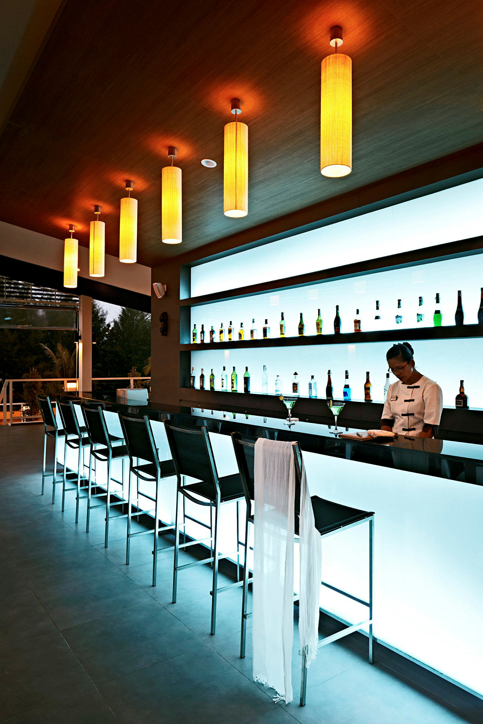 Moderne bar med en række høje stole og oplyst bagvæg med flasker. (AI-genereret tekst)