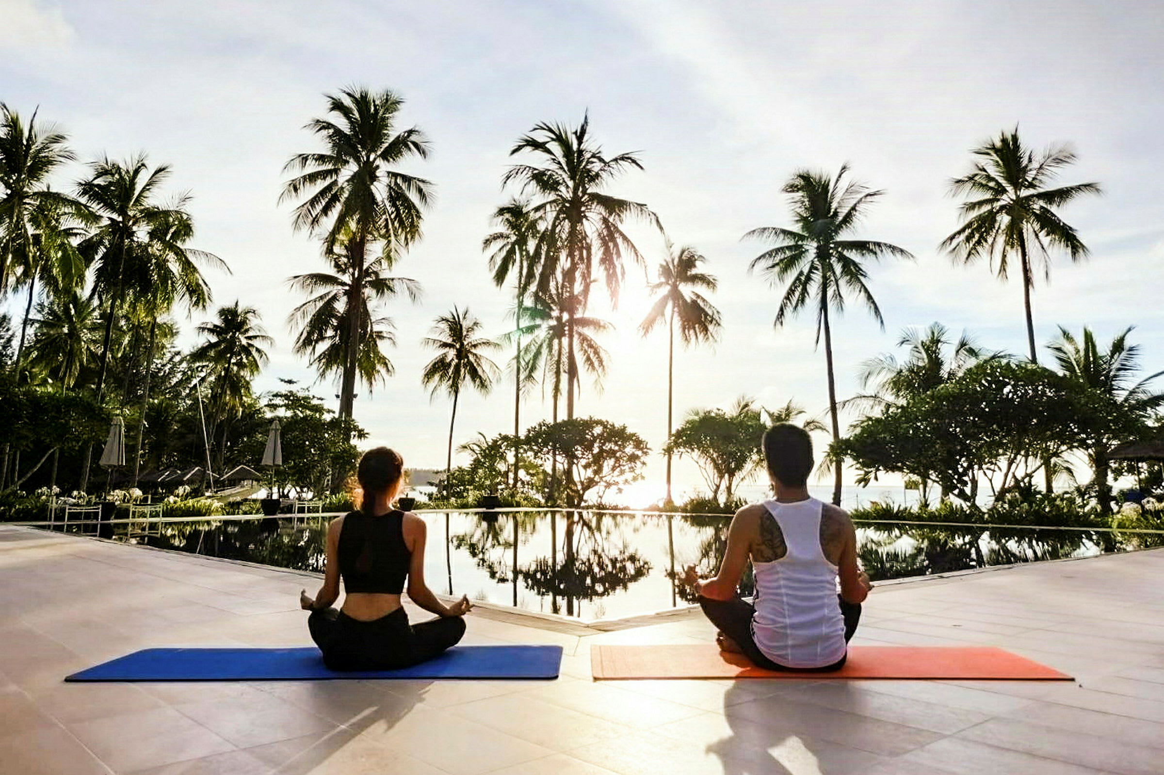 To personer sidder på yogamåtter ved en pool omgivet af palmer. (AI-genereret tekst)