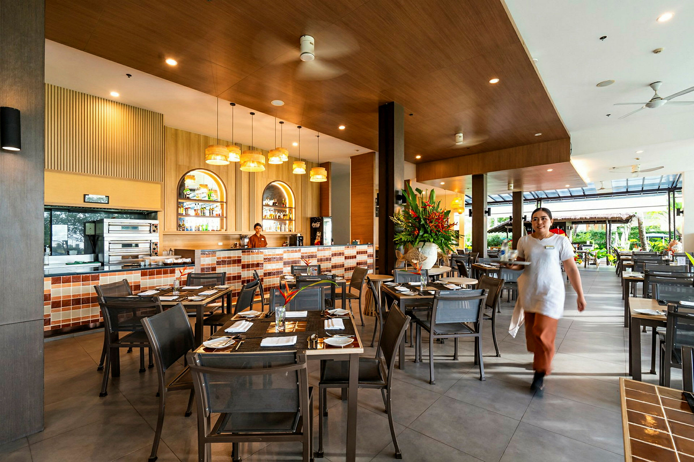 En restaurant med borde og stole; i baggrunden ses en bar med fliser og en medarbejder. En tjener går mod kameraet. (AI-genereret tekst)