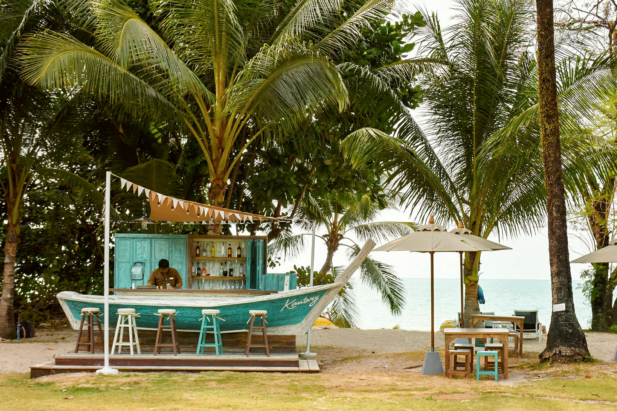 En strandbar bygget som en båd, omgivet af palmetræer og med udsigt over havet. Der er barstole foran baren og borde med parasoller ved siden af. (AI-genereret tekst)