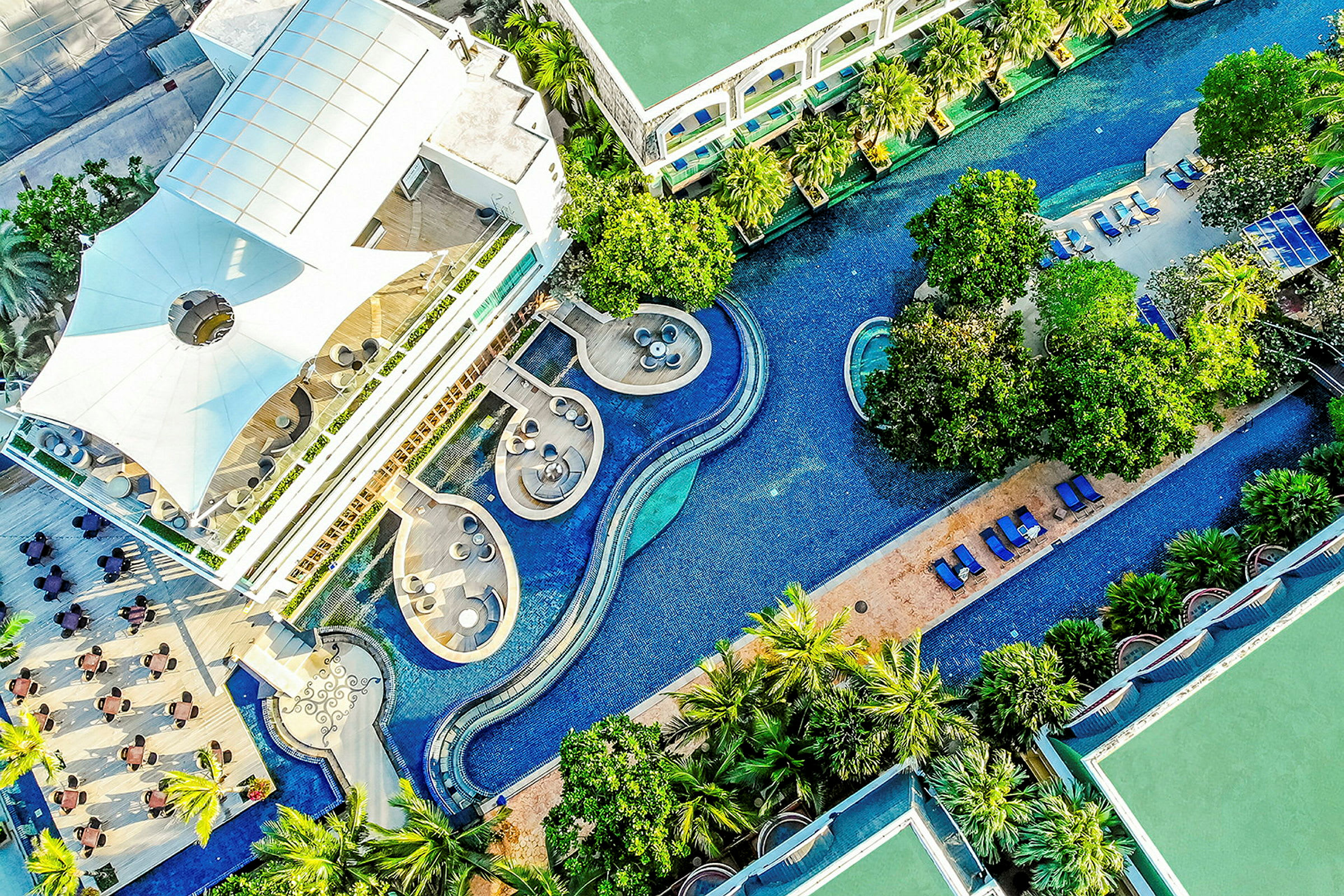 Luftfoto af en moderne bygning ved siden af en snoet swimmingpool omgivet af beplantning og liggestole. (AI-genereret tekst)